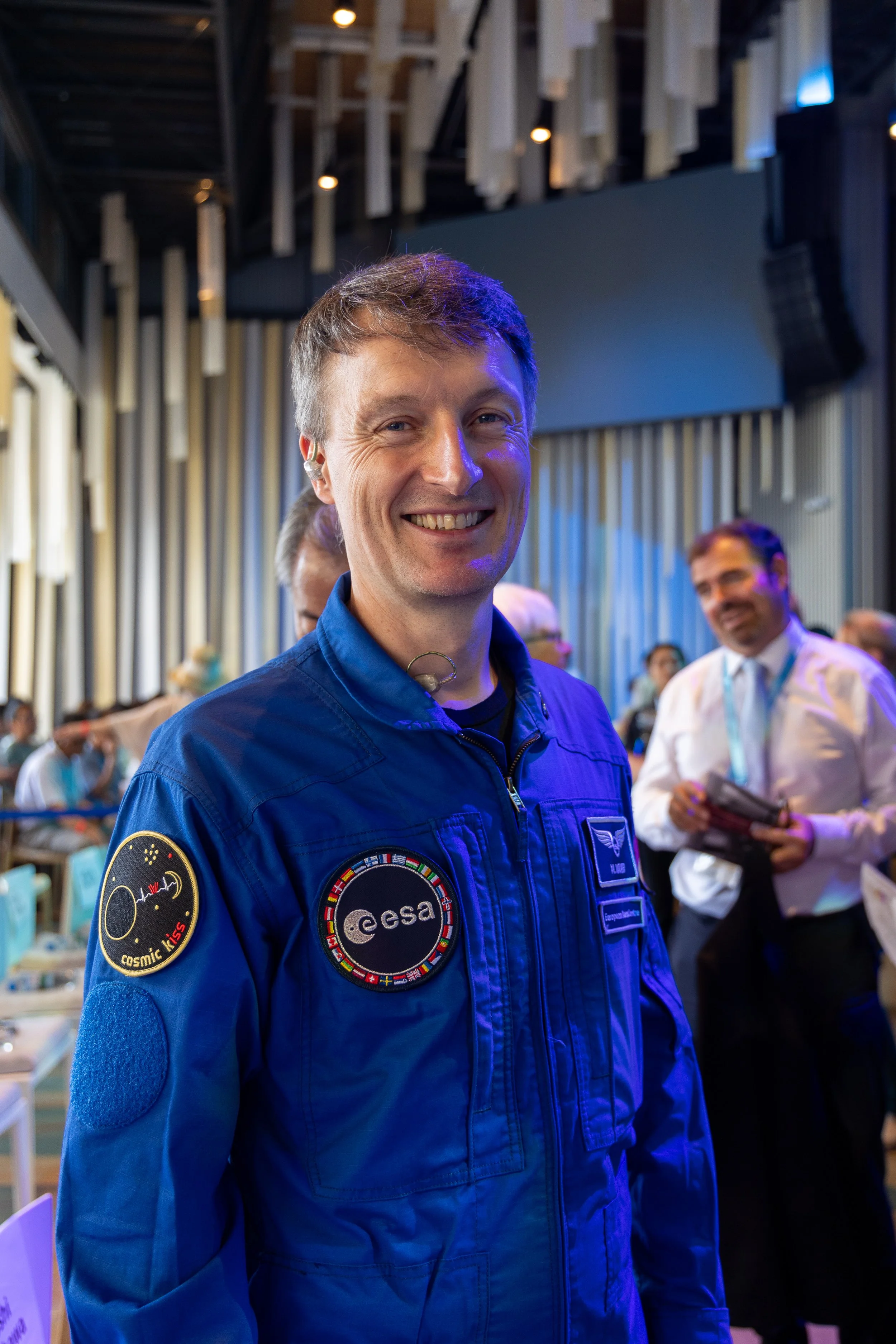 Un homme souriant en combinaison spatiale avec des logos de l'ESA, portant une décoration de badge, entouré de personnes dans une salle animée.