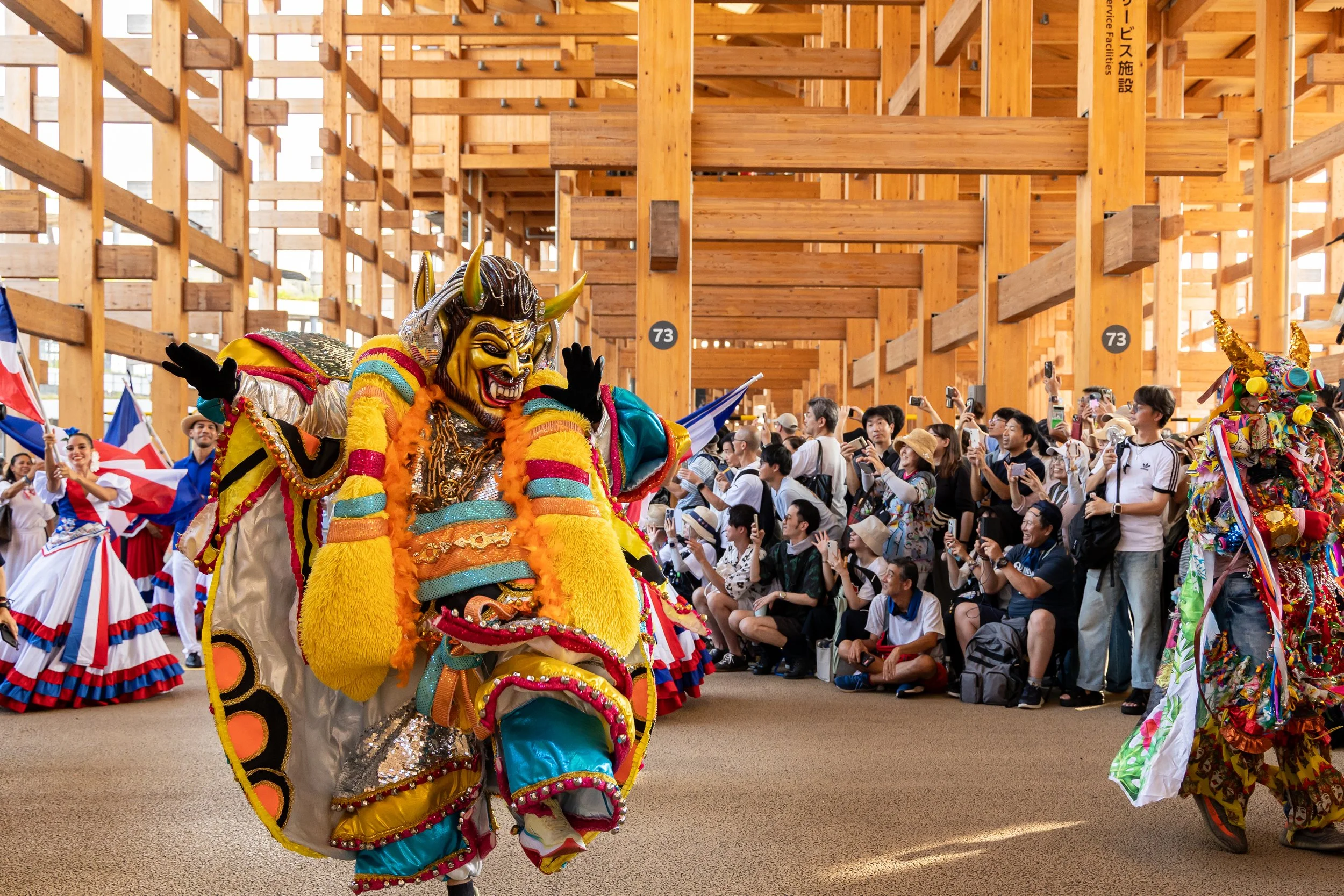 Un danseur portant un costume traditionnel coloré et un masque de demon participe à un festival ou parade. La scène est remplie de spectateurs qui prennent des photos, avec une structure en bois en arrière-plan.