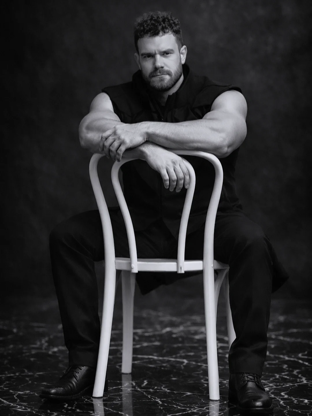 Black and white photo of a muscular man with short curly hair and beard, sitting on a chair and leaning on a backrest, looking serious.