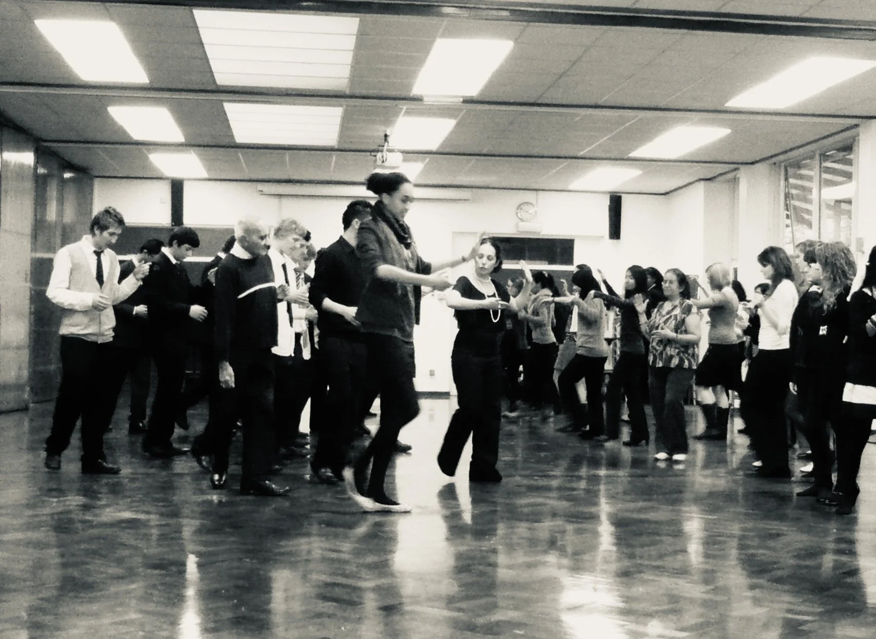 A group of people dancing together in a large room with a polished wooden floor, illuminated by fluorescent lights on the ceiling.