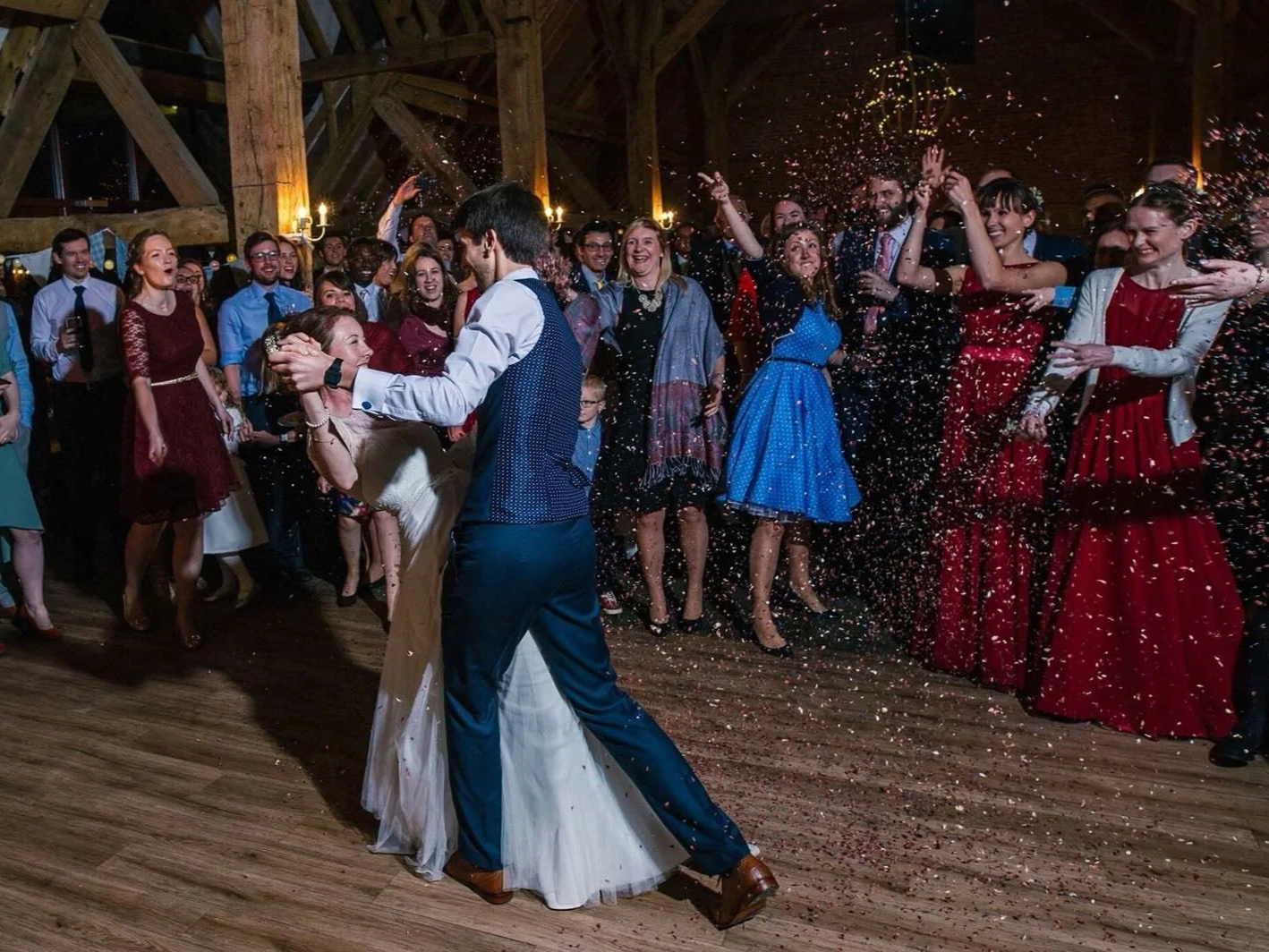 Couple dancing center stage at a wedding reception, surrounded by happy guests in a rustic barn setting, with confetti falling.