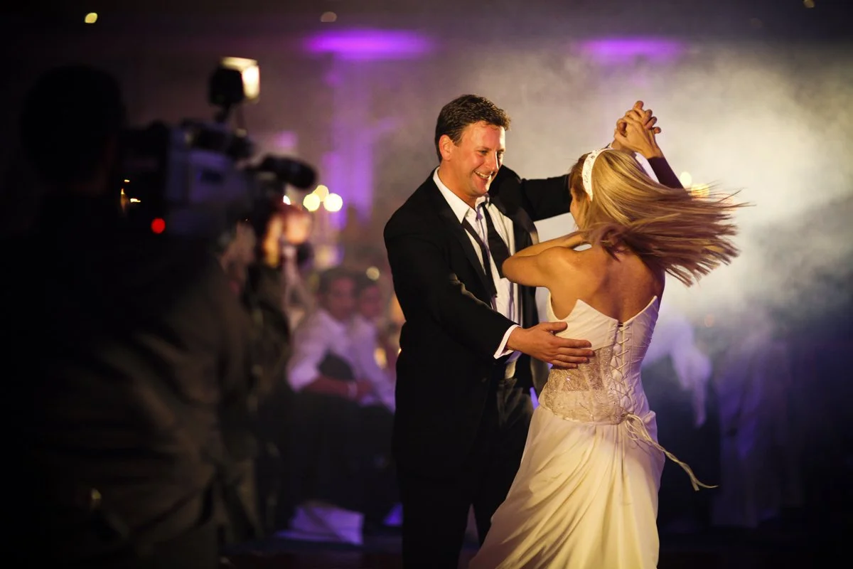 A couple dancing at a wedding reception, the man in a black suit and white shirt, and the woman in a white wedding dress with a ribbon and lace-up back, with guests and a photographer in the background.