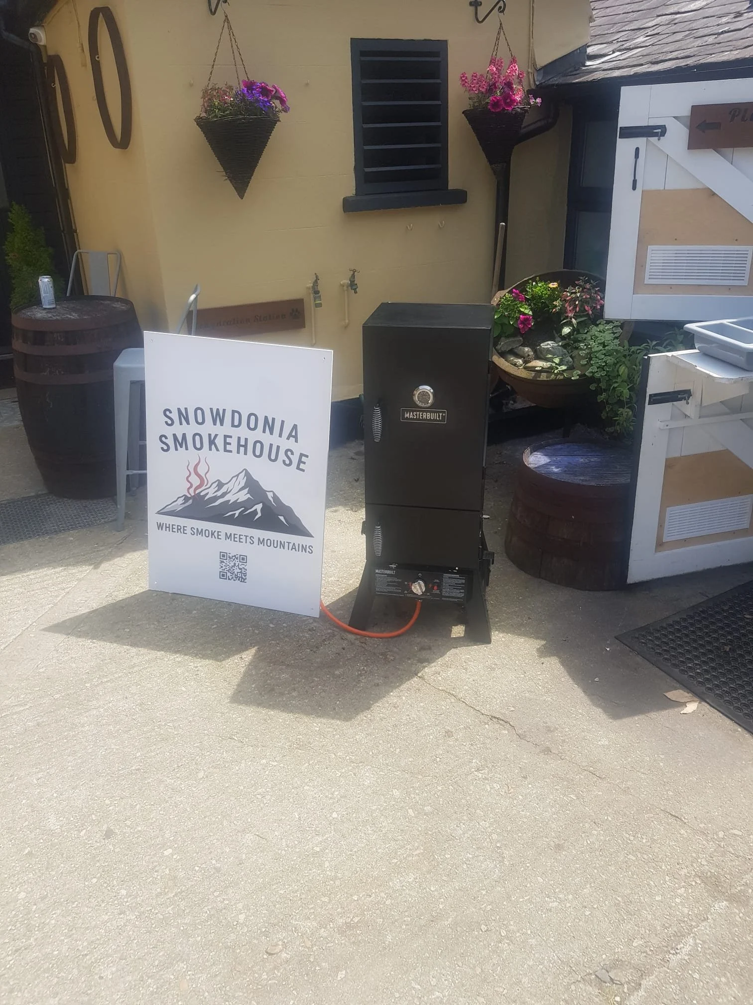 Outdoor scene featuring a sign for Snowdonia Smokehouse with mountain and smoke graphic, a black smoker grill, hanging flower baskets, potted flowers, a small table, and various decorative elements.