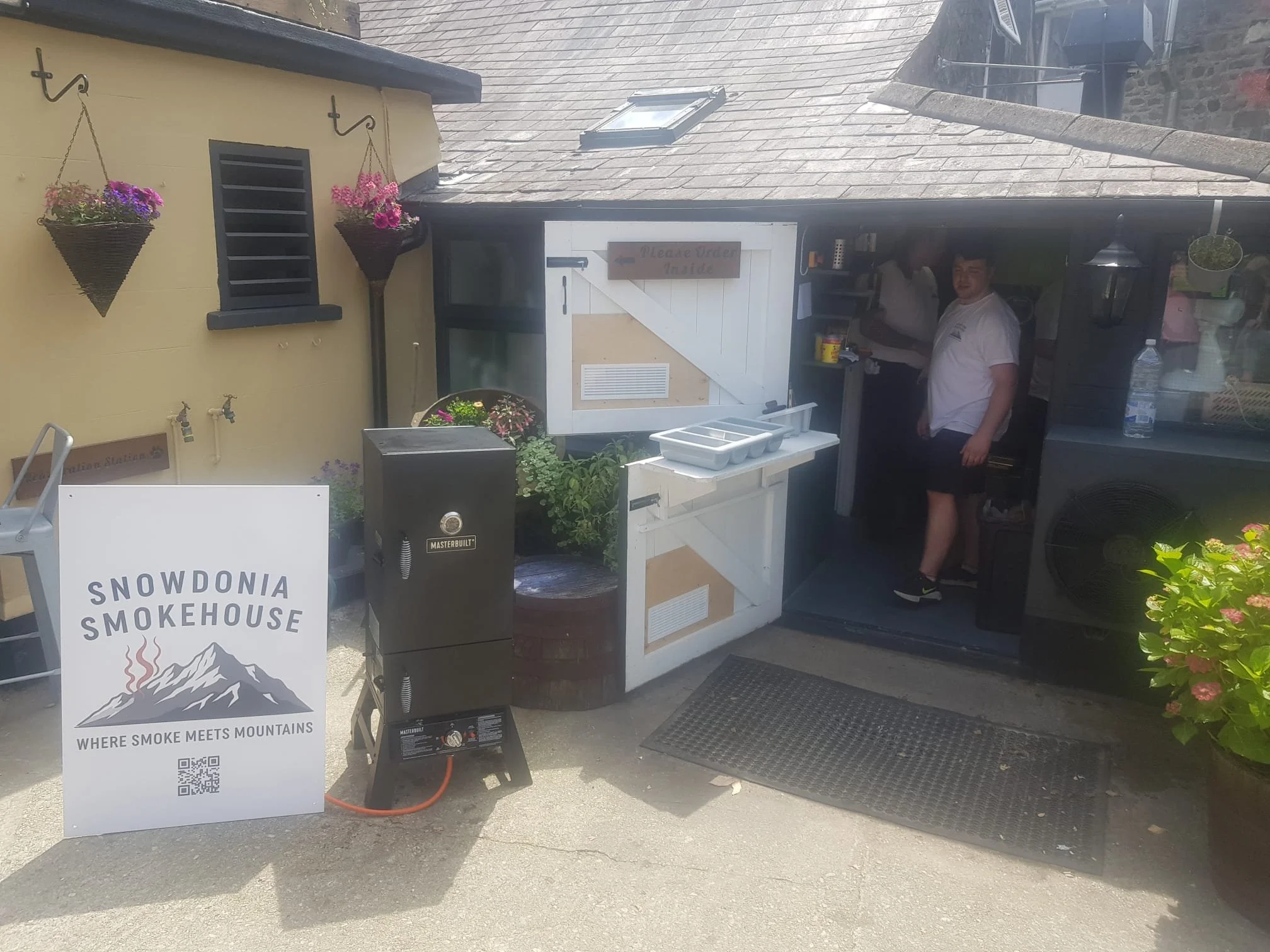 A small smokehouse with a sign reading "Snowdonia Smokehouse" and the slogan "Where smoke meets mountains." There are potted plants, hanging flower baskets, and a Masterbuilt smoker grill outside. Two people are inside the smokehouse, one at the coun