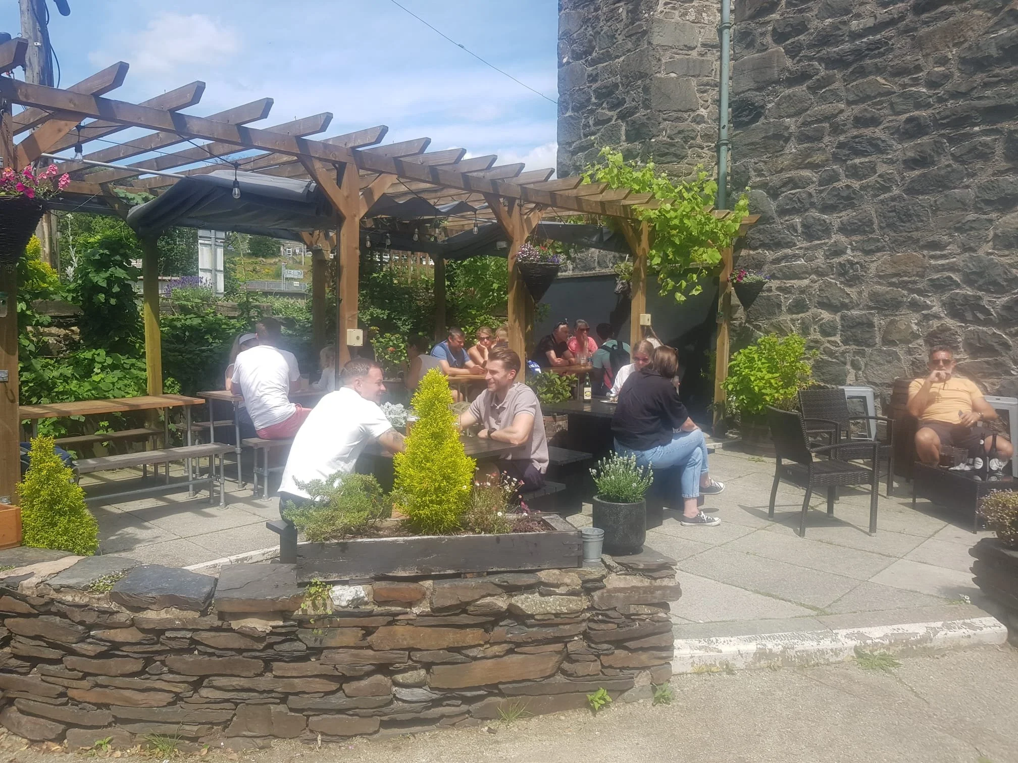 Outdoor patio with people sitting at tables under a wooden pergola with hanging flower baskets and string lights, surrounded by greenery and stone walls.