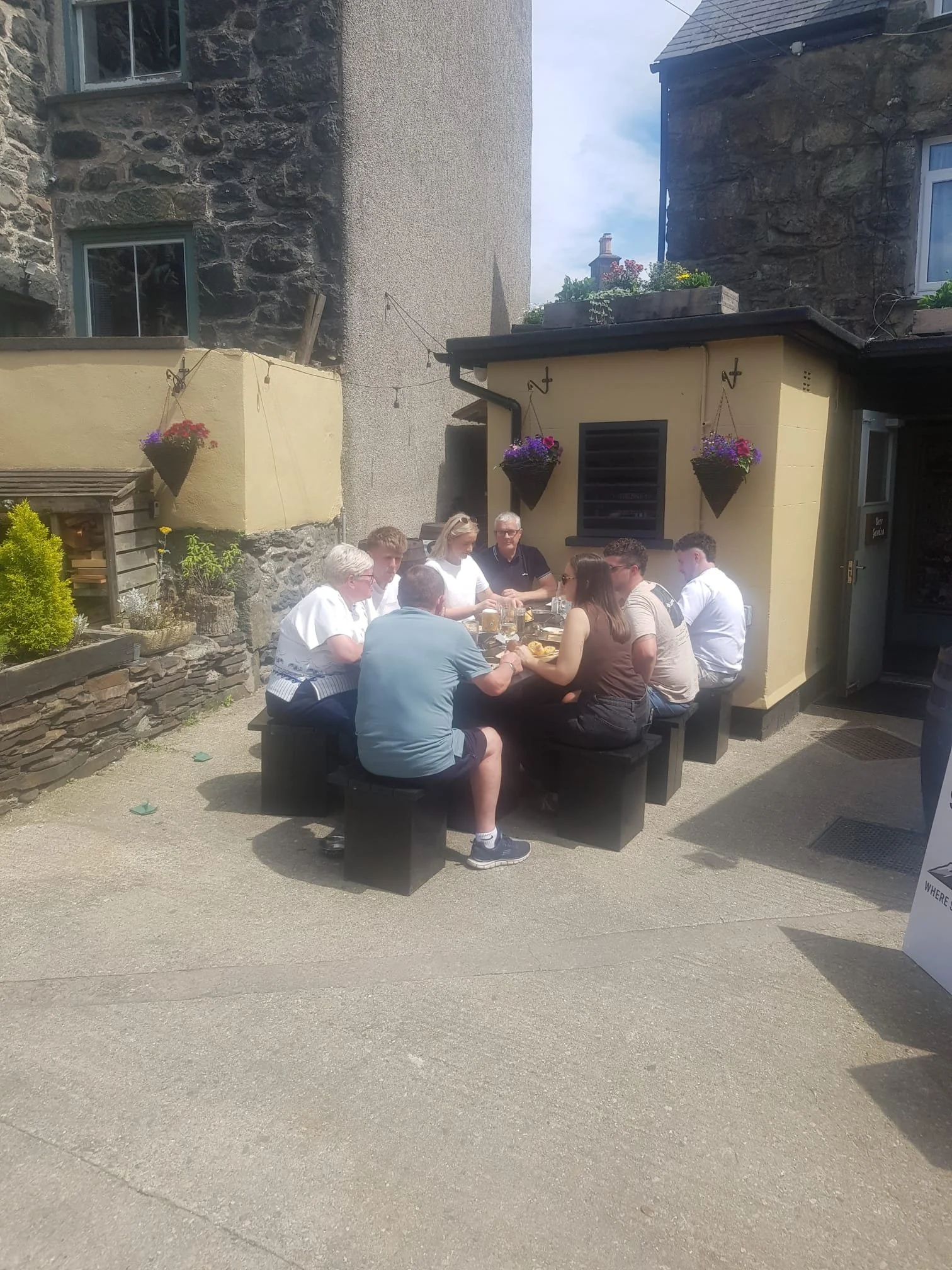 Group of people sitting around a picnic table outside a building, enjoying food and drinks on a sunny day.