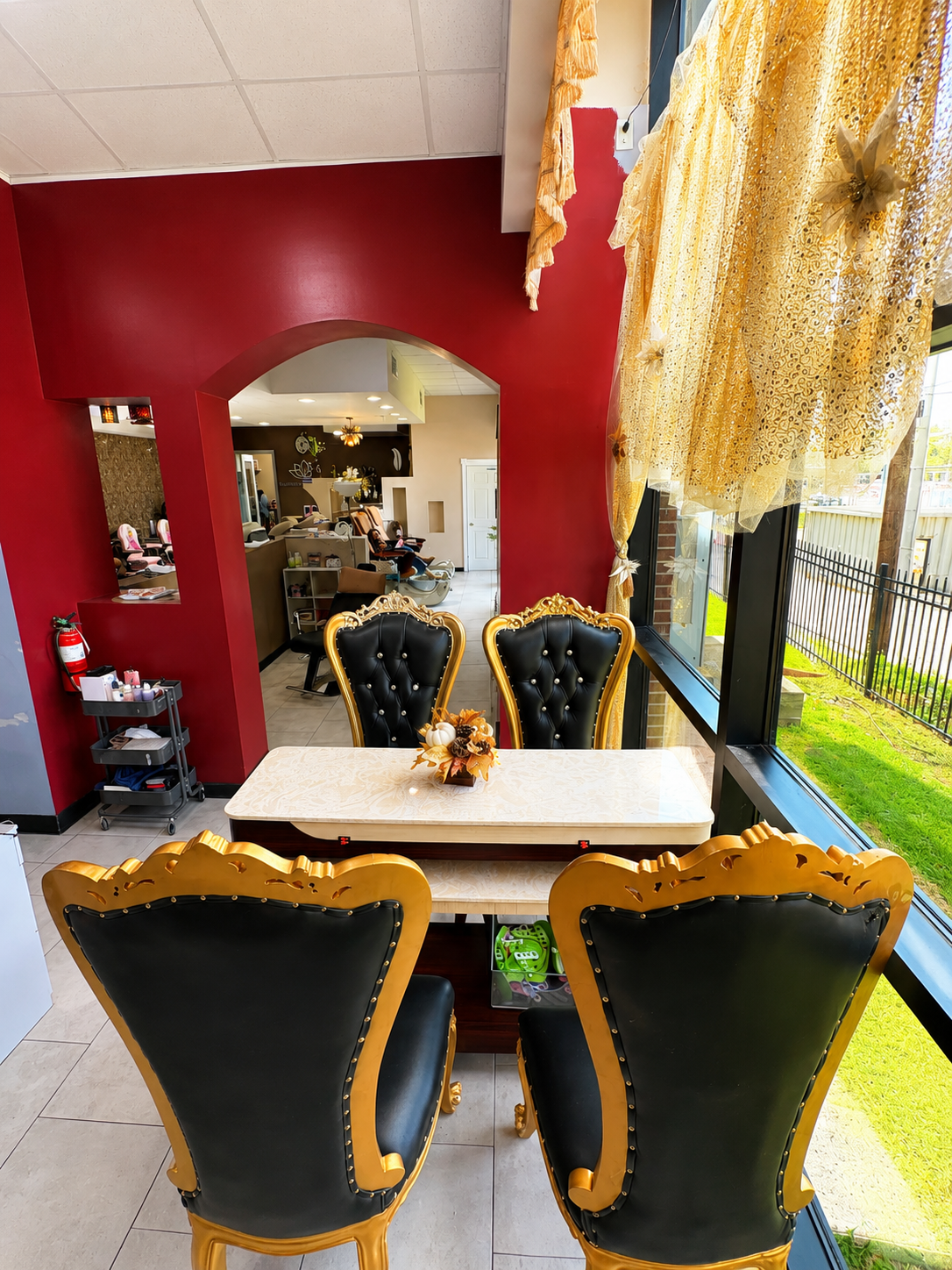 Interior of a nail salon featuring a small table and four ornate black and gold chairs near a large window with yellow lace curtains, overlooking a fenced yard with grass.