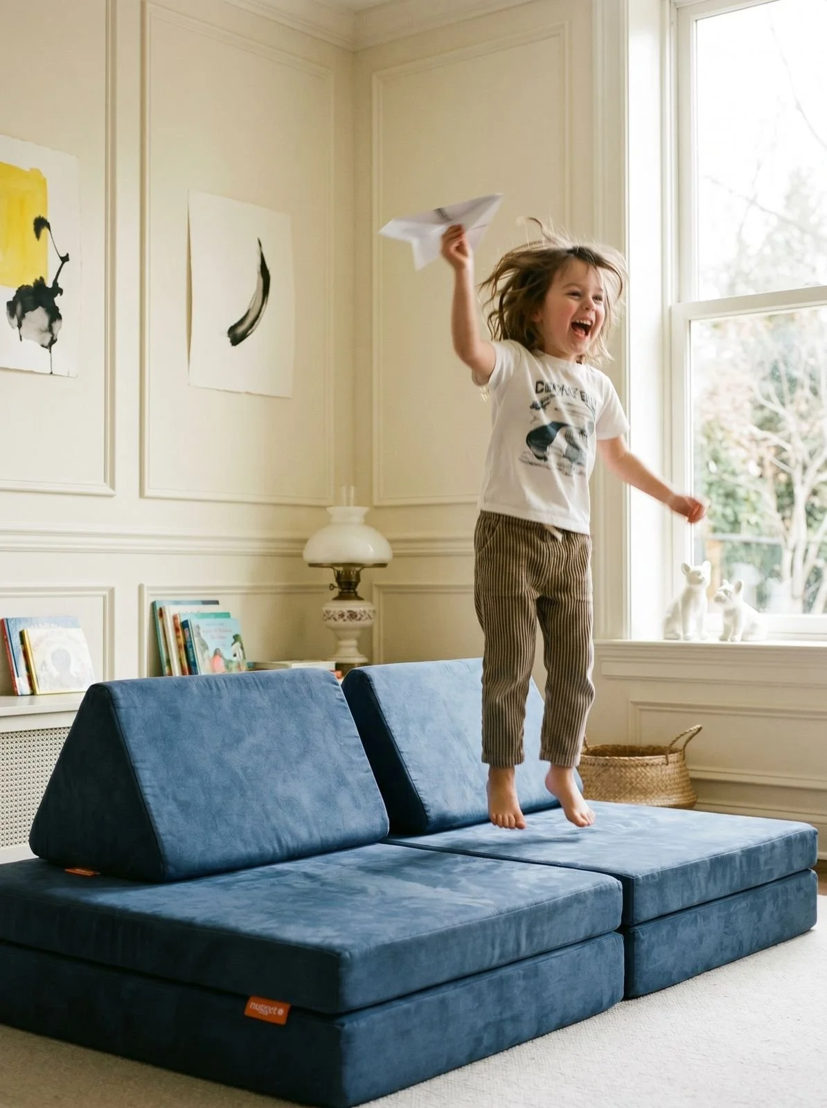 Child jumping on a blue couch while playing with a paper airplane in a bright living room.