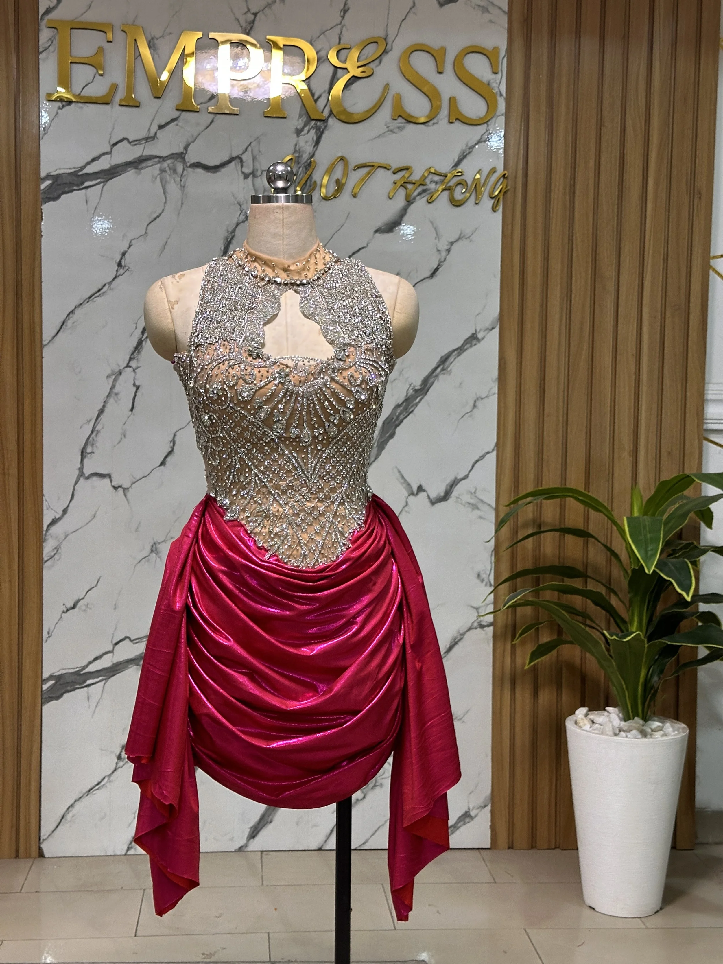 A mannequin dressed in a glamorous pink gown with a heavily embellished silver and rhinestone bodice, displayed in front of a backdrop with the words 'Empress Hot Hong' and a marble wall. There is also a potted green plant on the right side.