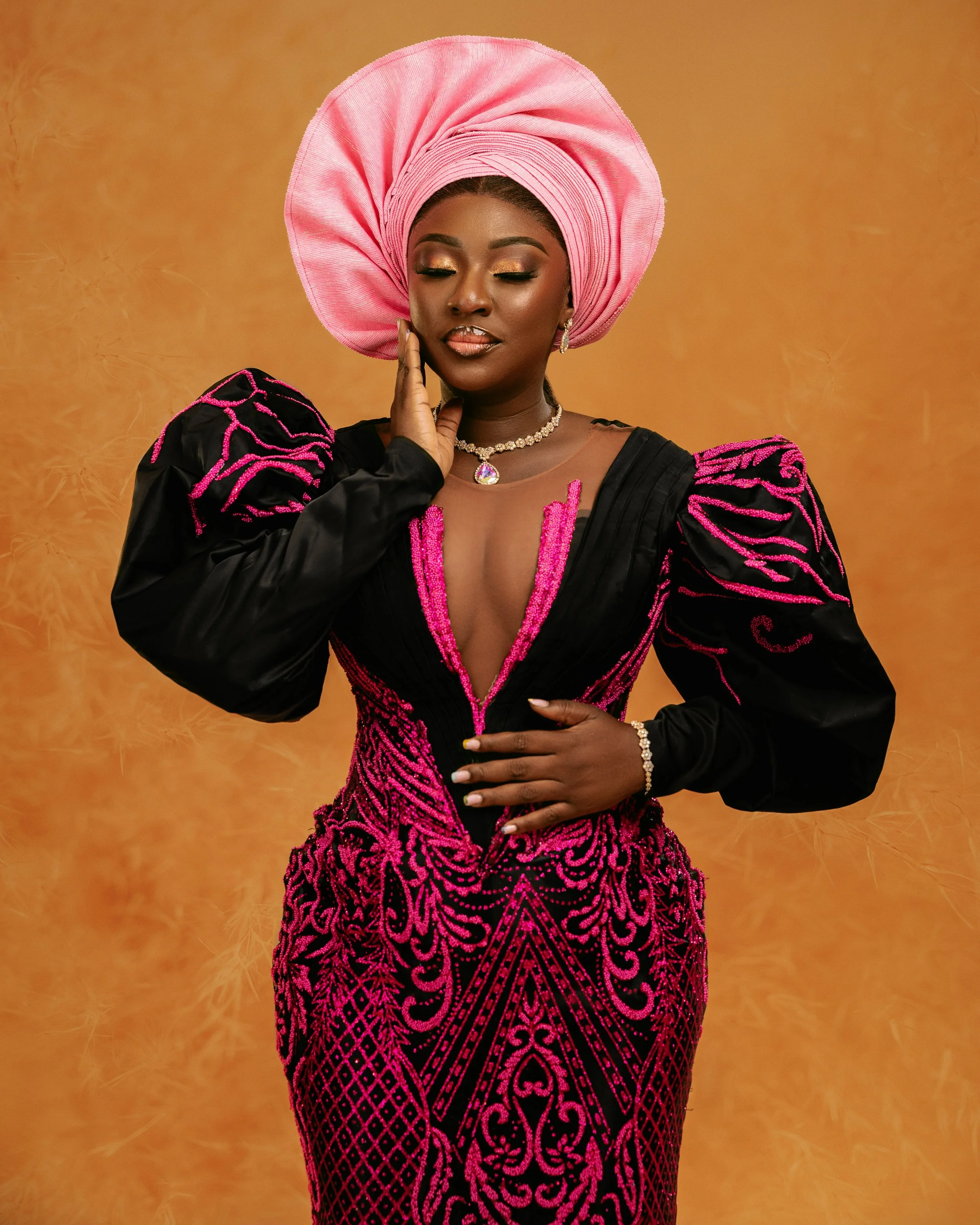 A woman wearing traditional Nigerian attire, including a pink gele (headwrap) and a black and pink gele. She is dressed in a black dress with pink embroidery, accessorized with jewelry, and stands against a warm yellow background.