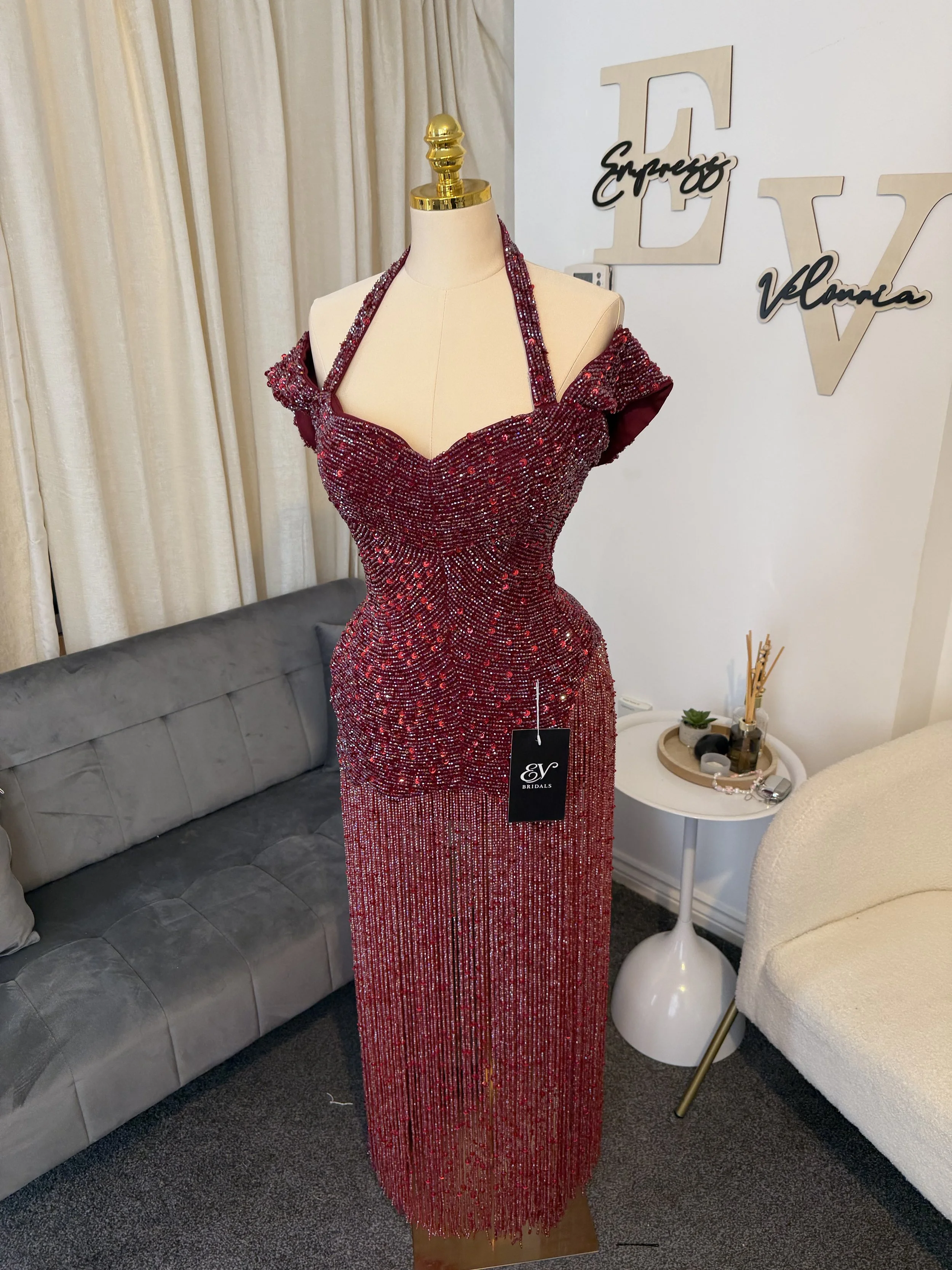 A mannequin wearing a red, sequined evening gown with off-shoulder and halter straps, displayed inside a boutique. The boutique has a gray sofa, a white side table with decorative items, and a wall with wooden letters and the words "Express Velluma."