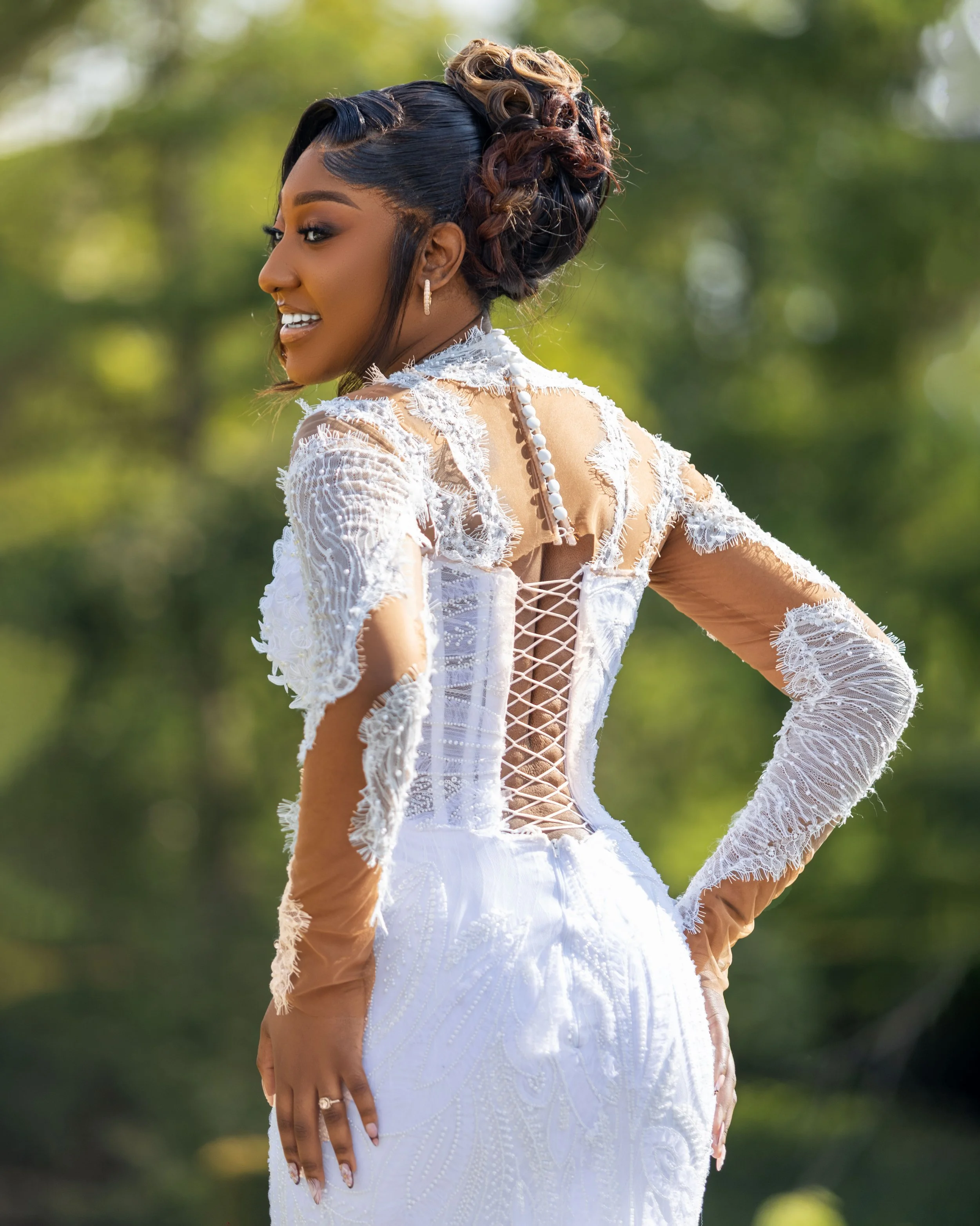 A woman with a styled updo hairstyle, wearing a white lace dress with sheer sleeves, standing outdoors with trees in the background.