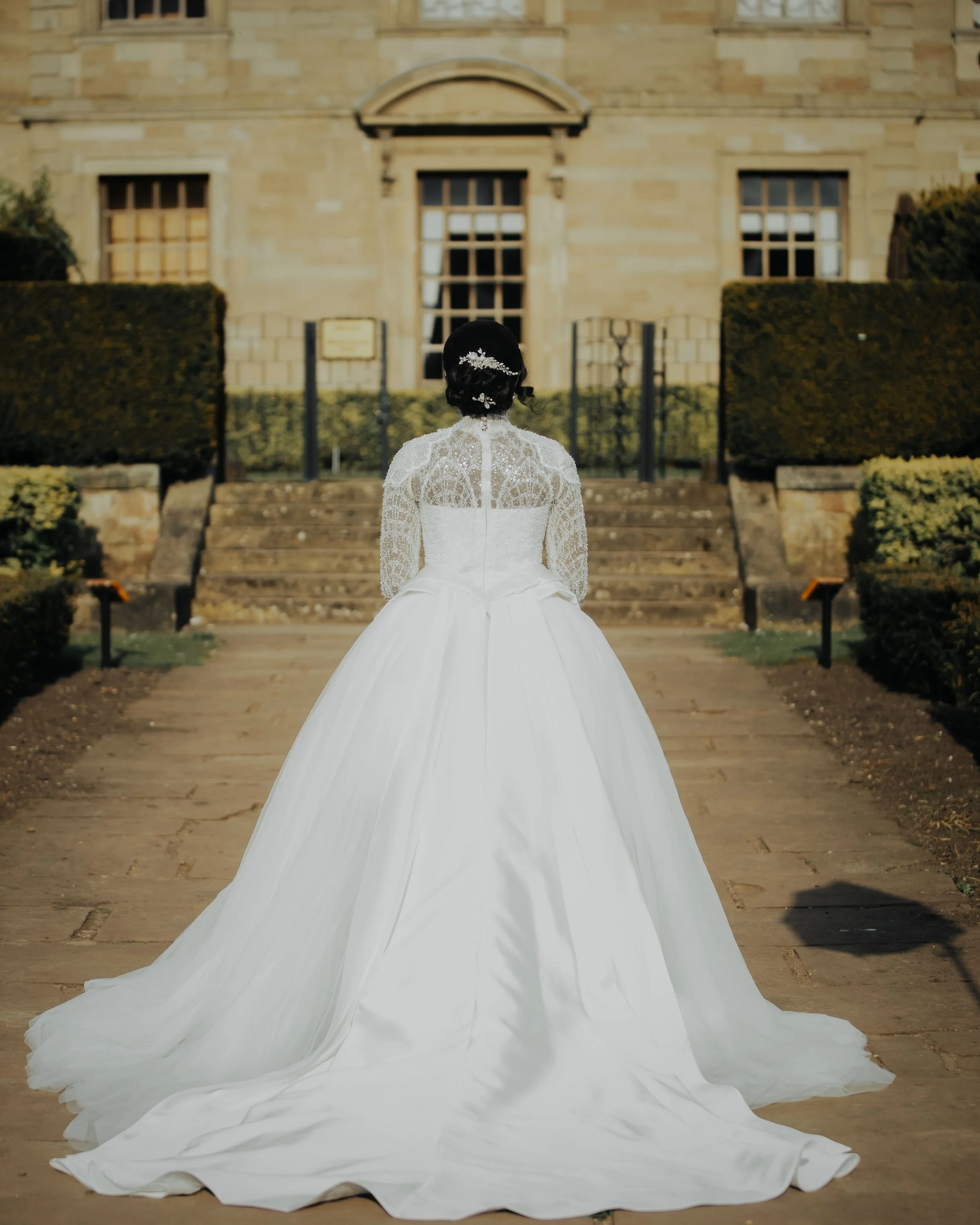 Back view of a bride in a white wedding dress with lace upper part and long flowing skirt, sitting on stairs outside a historic building with stone walls, large windows, and trimmed hedges.