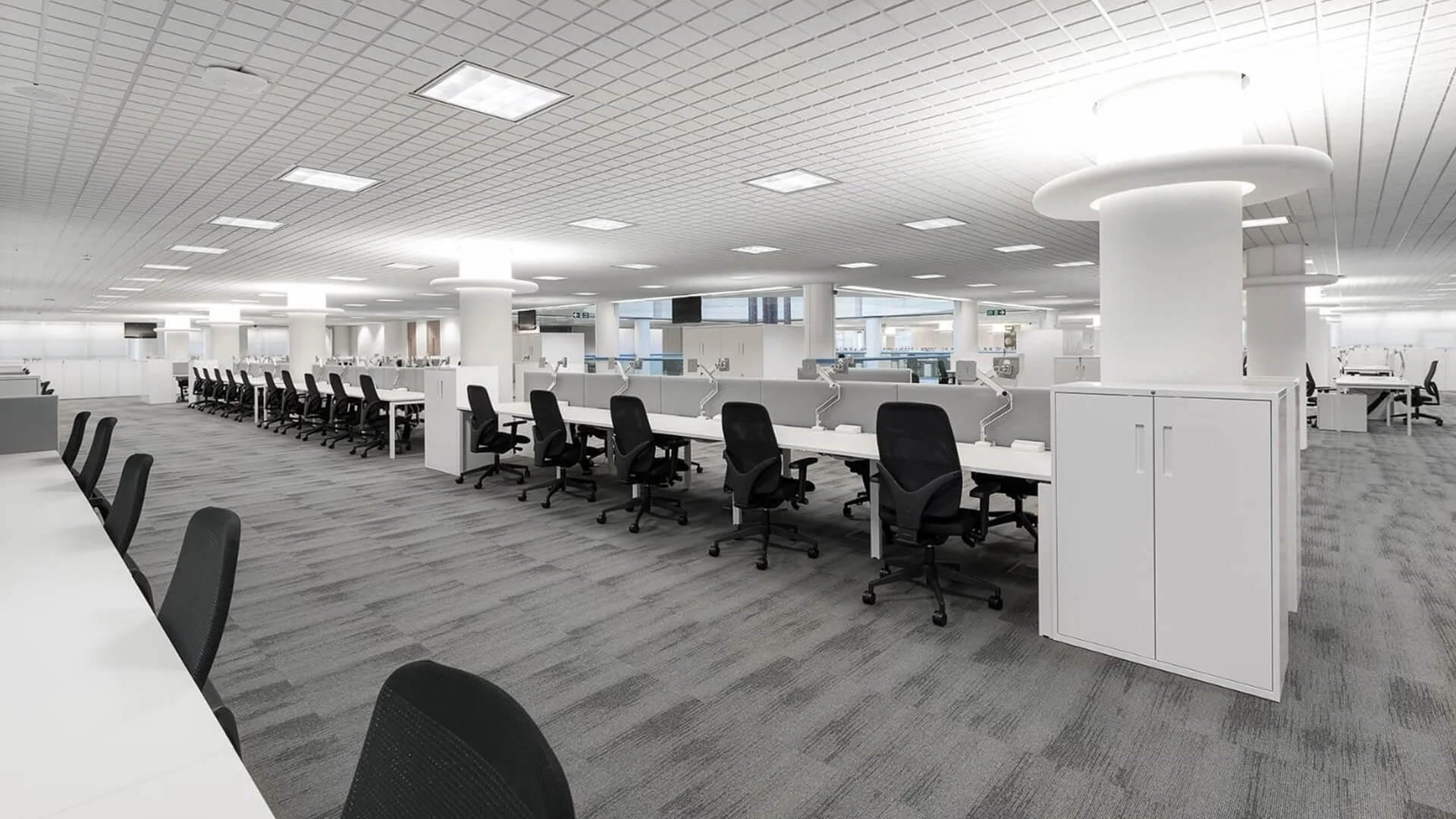 A modern, open-plan office with white desks and black chairs, partitioned workspace areas, and large windows providing natural light. Ceiling has rectangular lighting panels and circular light fixtures.