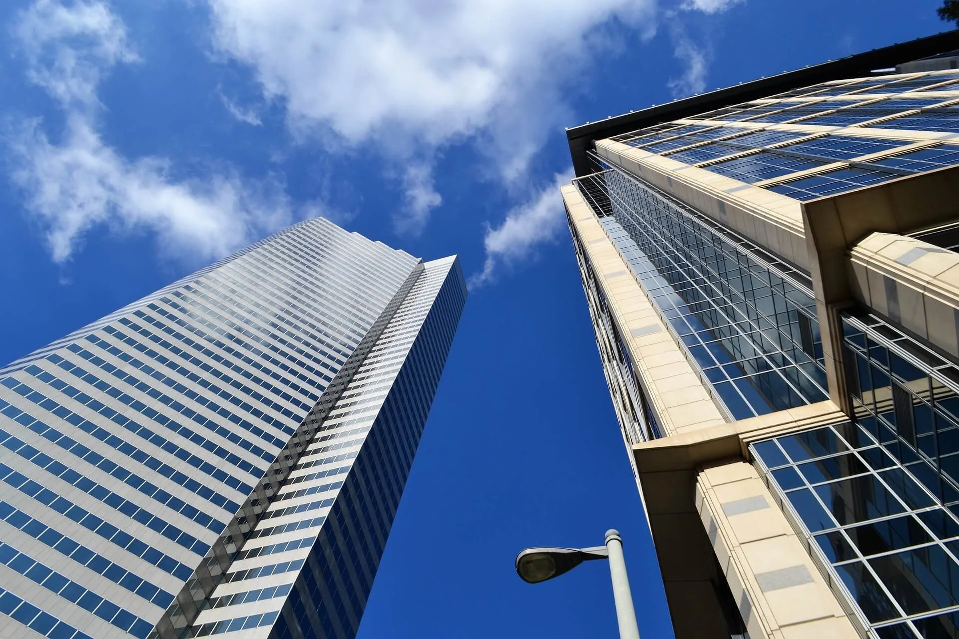 View of tall modern skyscrapers with glass facades under a blue sky with some clouds, including a streetlamp in the lower part of the image.