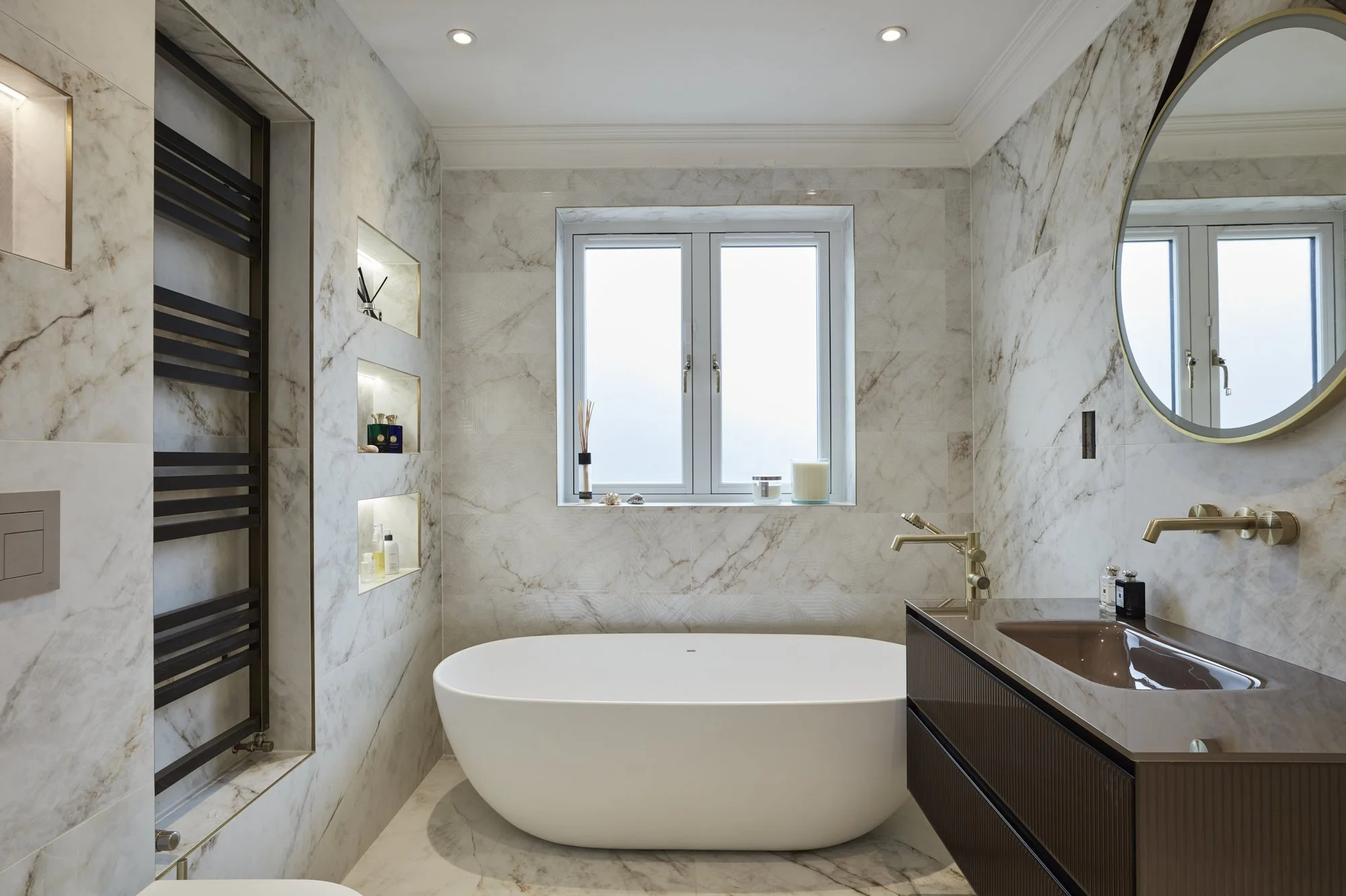 Modern bathroom with marble walls, a freestanding bathtub, a window, a black sink with a brown basin, and built-in shelves with toiletries.