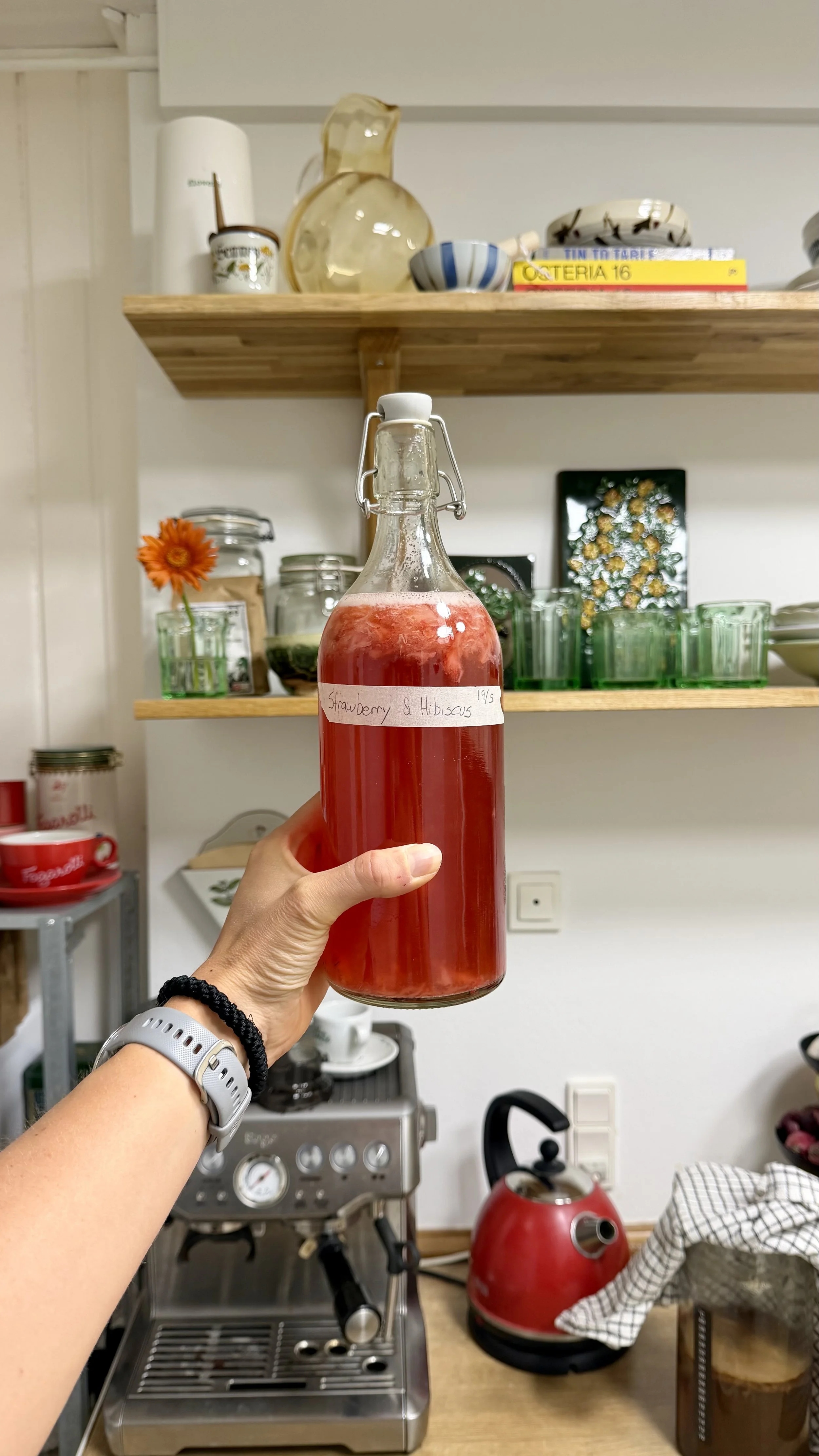 A hand holding a glass bottle with a red liquid and a white label reading 'Strawberry & Hibiscus' in a kitchen. The background has shelves with dishes, a coffee machine, a red kettle, and other kitchen items.