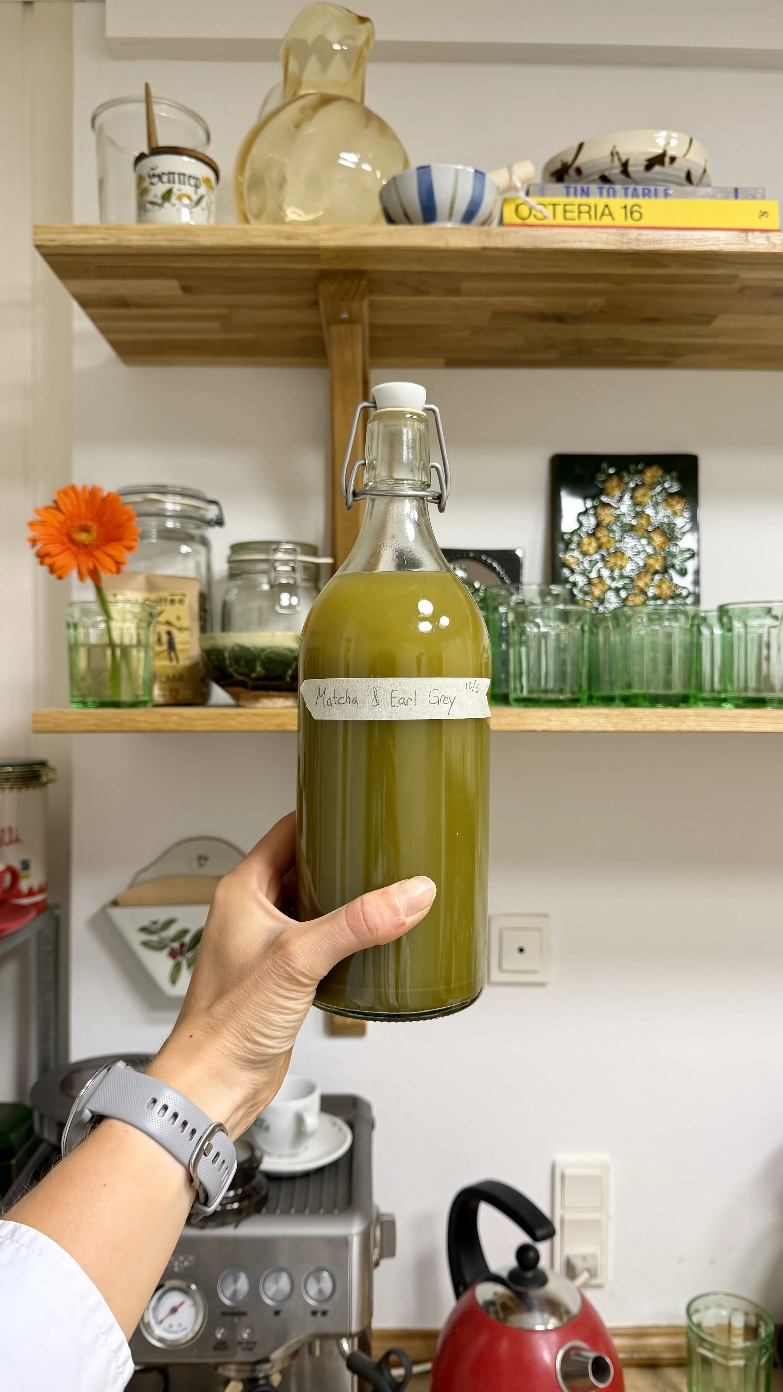Hand holding a glass bottle labeled 'Matcha & Earl Grey' in a kitchen. Background includes shelves with green glasses, jars, a flower, and kitchen appliances.