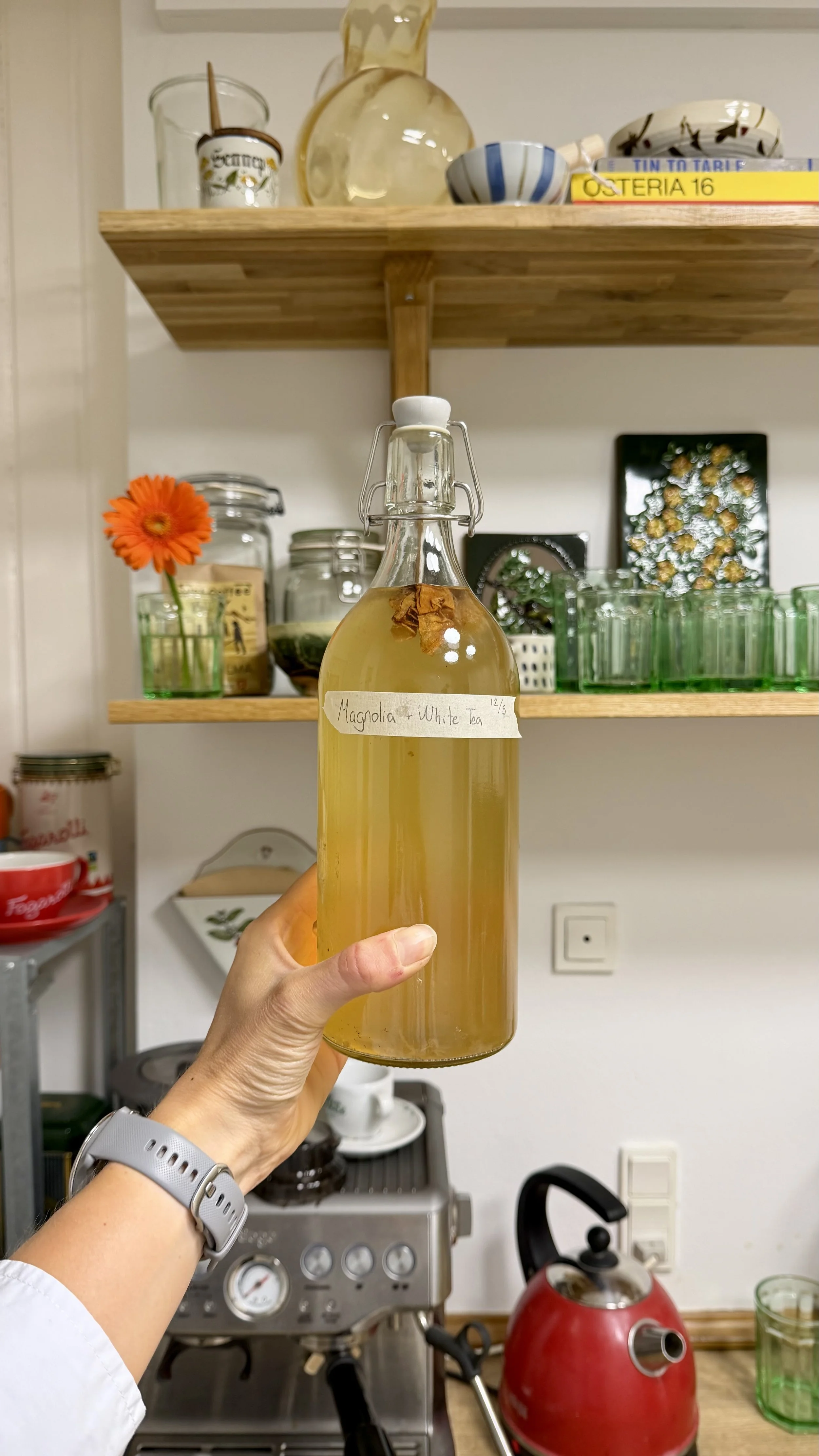 A hand holding a glass bottle labeled "Magnolia + White Tea" in a kitchen. In the background, there are shelves with kitchen items, a coffee machine, a red kettle, and various cups and containers.