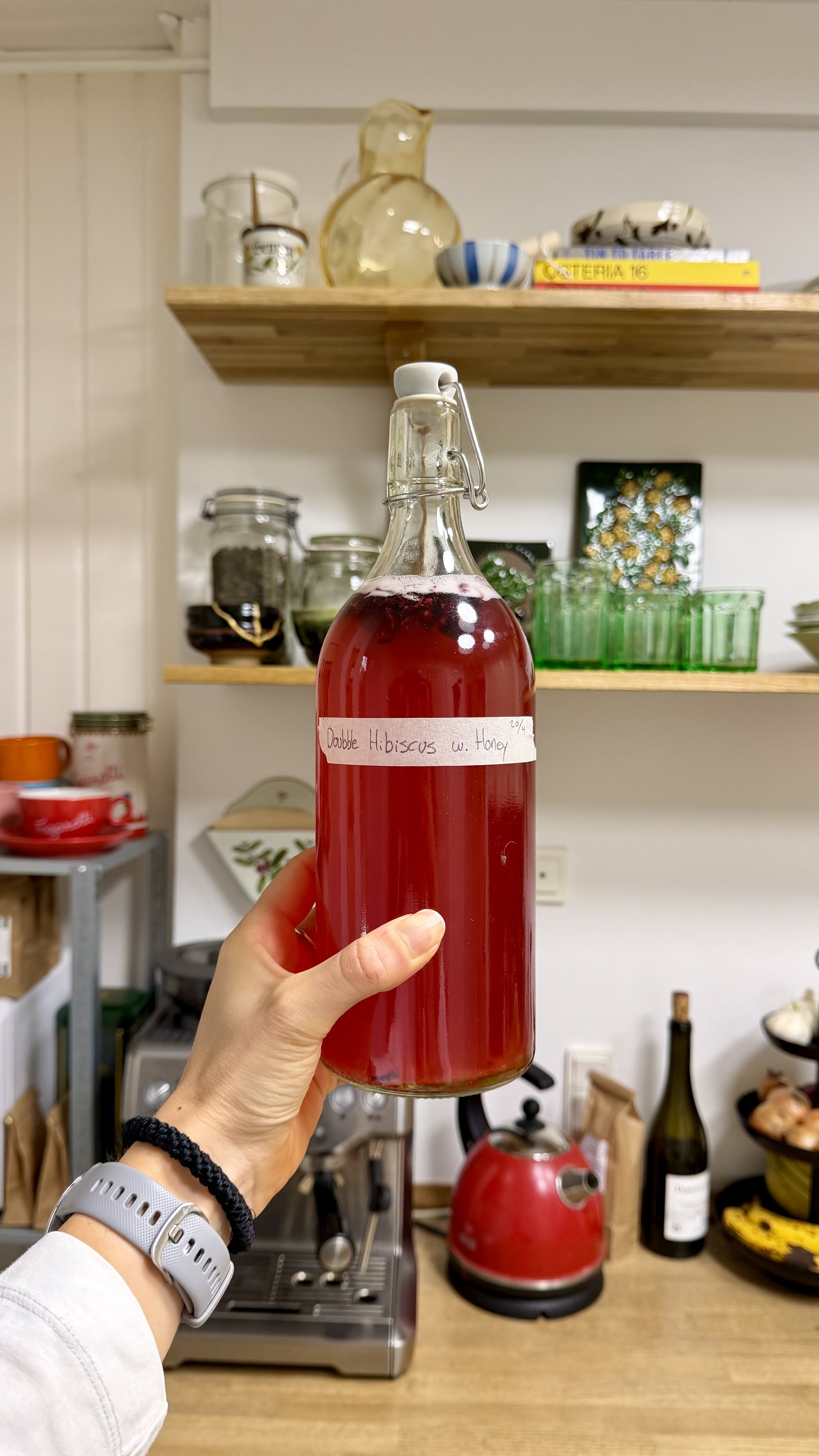 A person holding a glass bottle labeled "Dapple Hibiscus w. Honey" filled with a red beverage in a kitchen setting with shelves, jars, a coffee machine, a red kettle, wine bottles, and other kitchen items in the background.