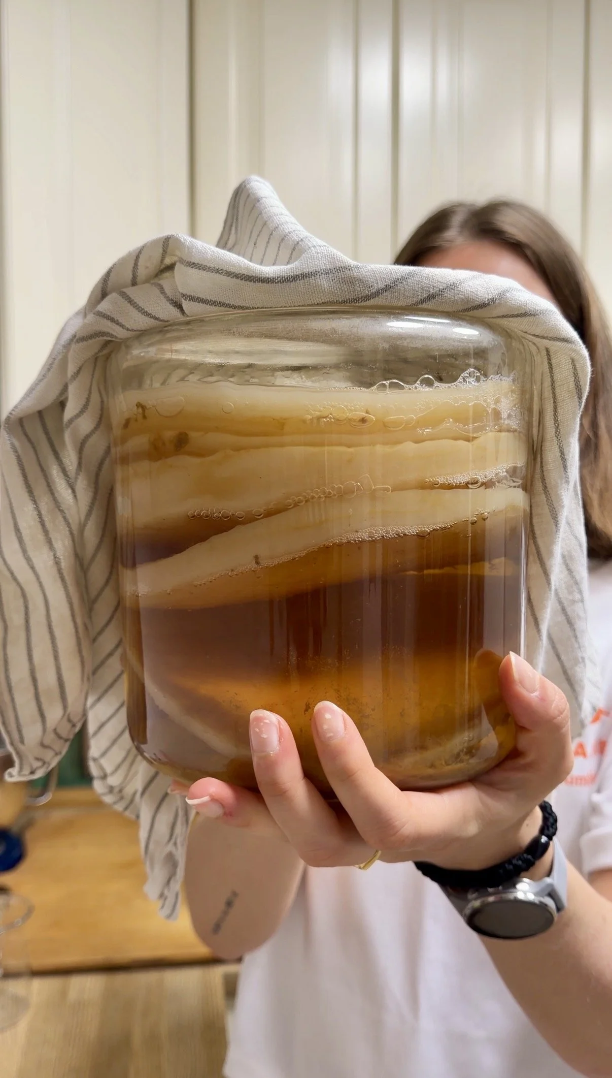 Person holding a large jar of kombucha tea with visible layers of SCOBY inside, partially covering their face.