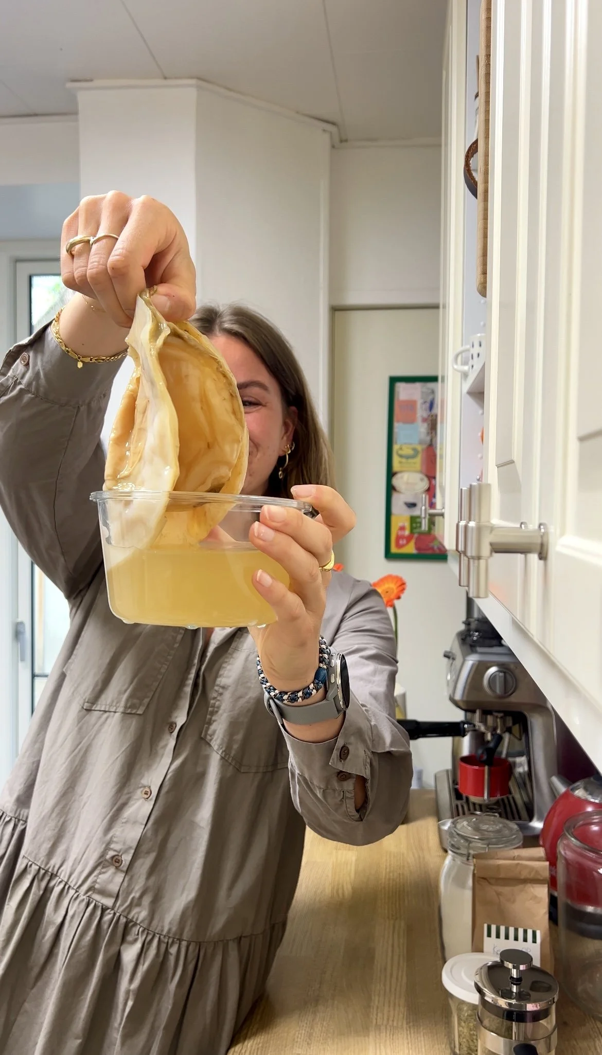 A woman with long hair and jewelry, wearing a beige dress, is holding a jar of honey and dipping a large chunk of beeswax or honeycomb into it in a kitchen.
