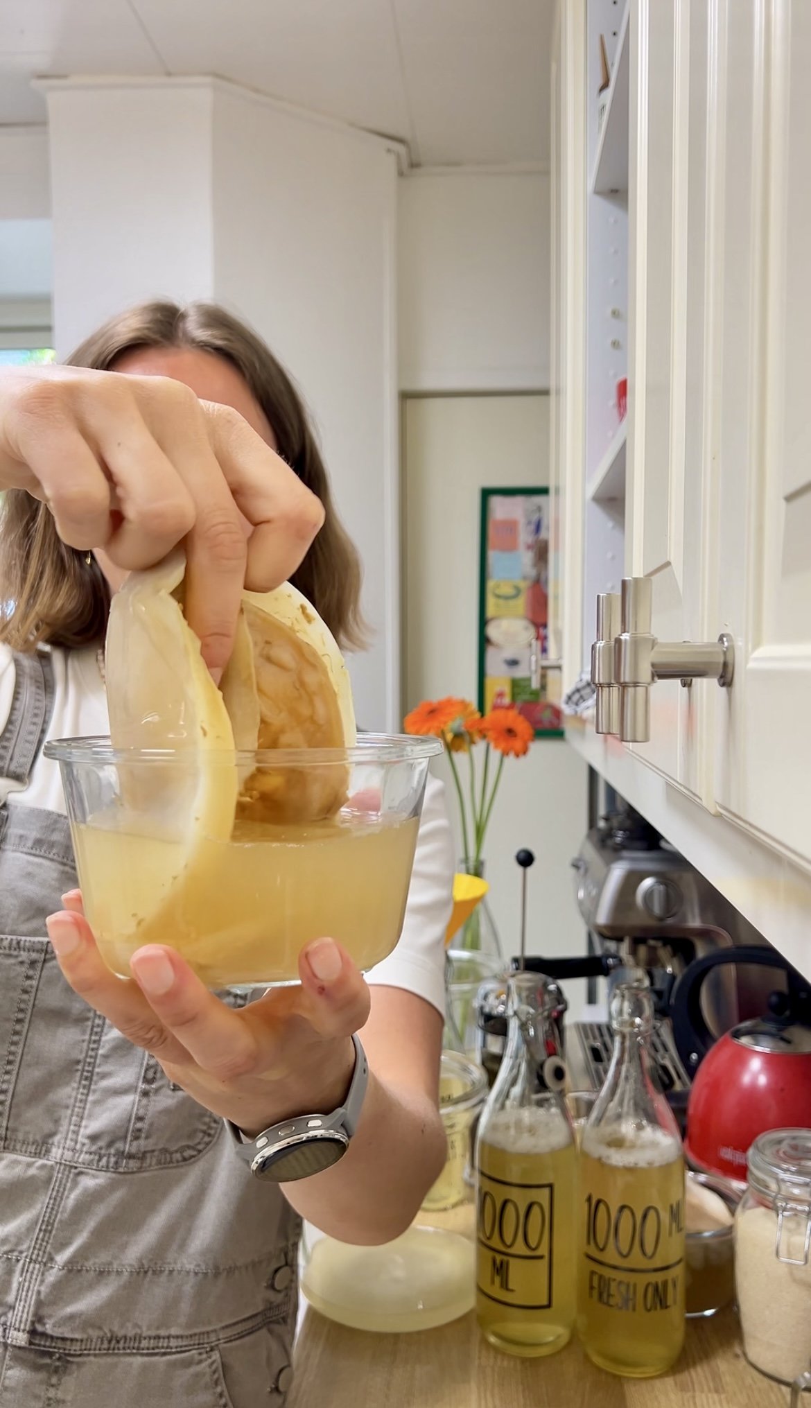 Person pouring liquid from a lemon into a glass container on a kitchen countertop, with various bottles and kitchen appliances in the background.