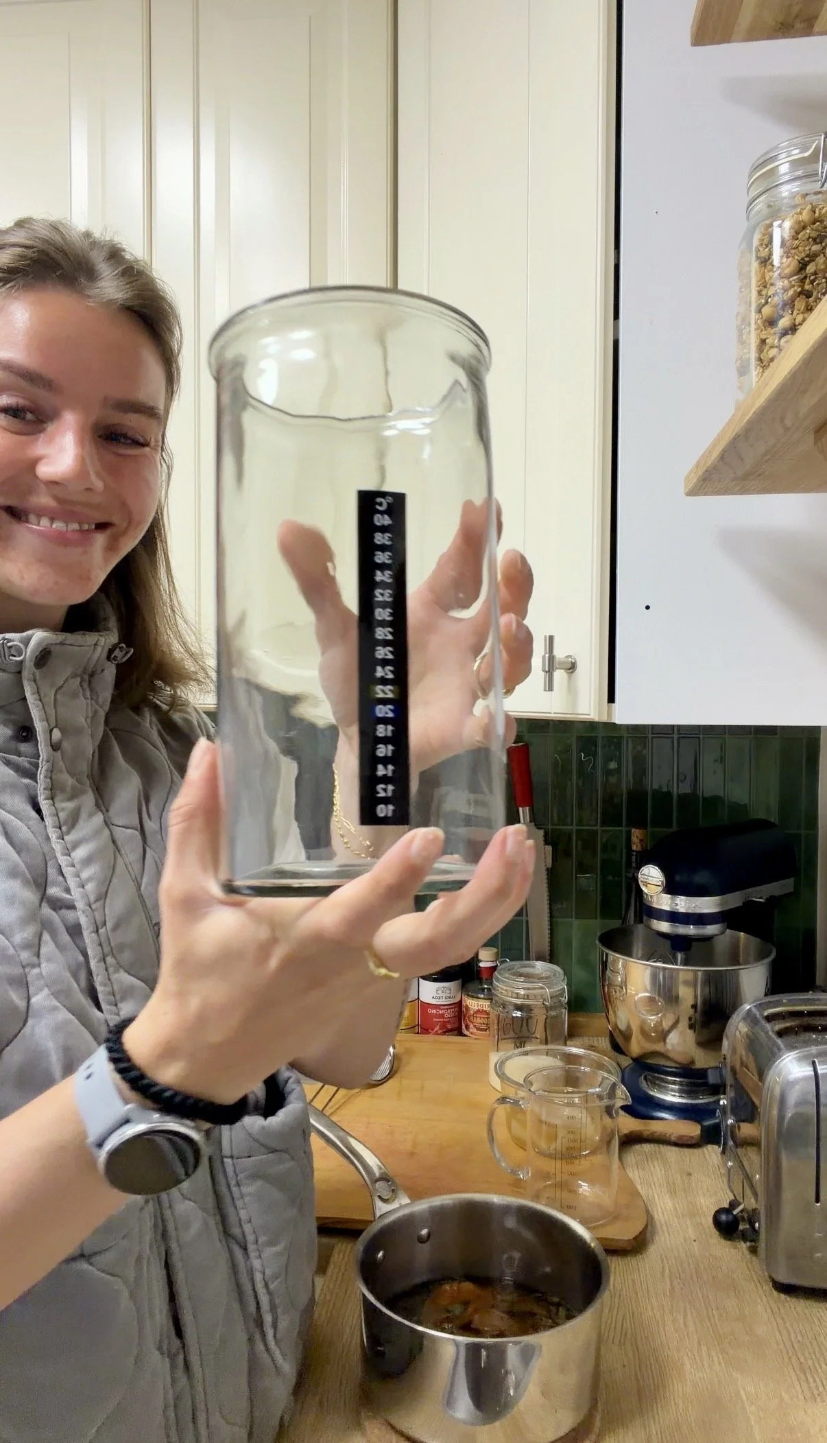 A woman in a kitchen holding a clear glass jar with a black and white measuring scale on it, smiling. The kitchen has white cabinets, various jars, a stand mixer, a pot with dark liquid, and a knife on the countertop.