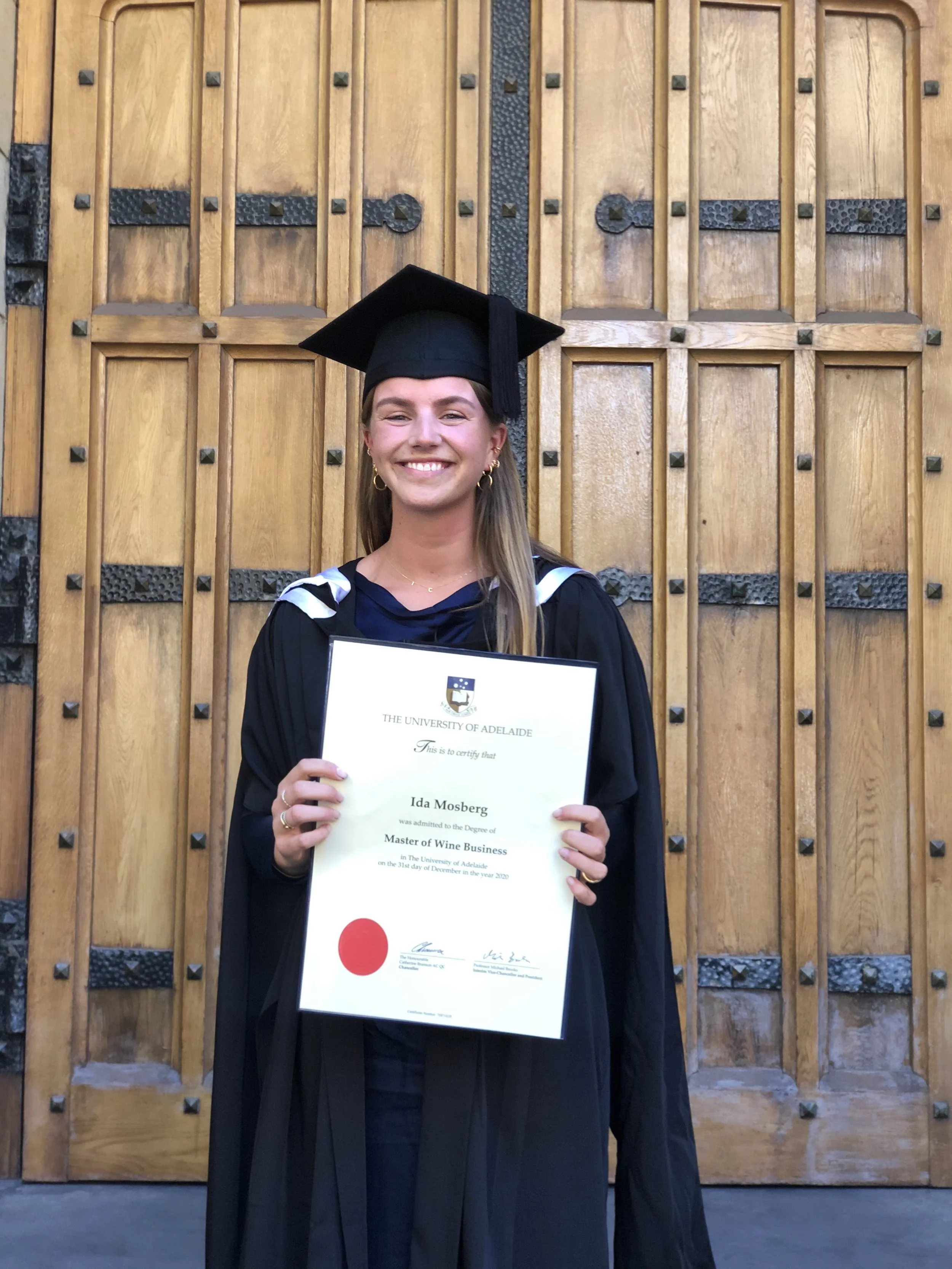 A young woman in a graduation gown holding a diploma in front of a wooden door.