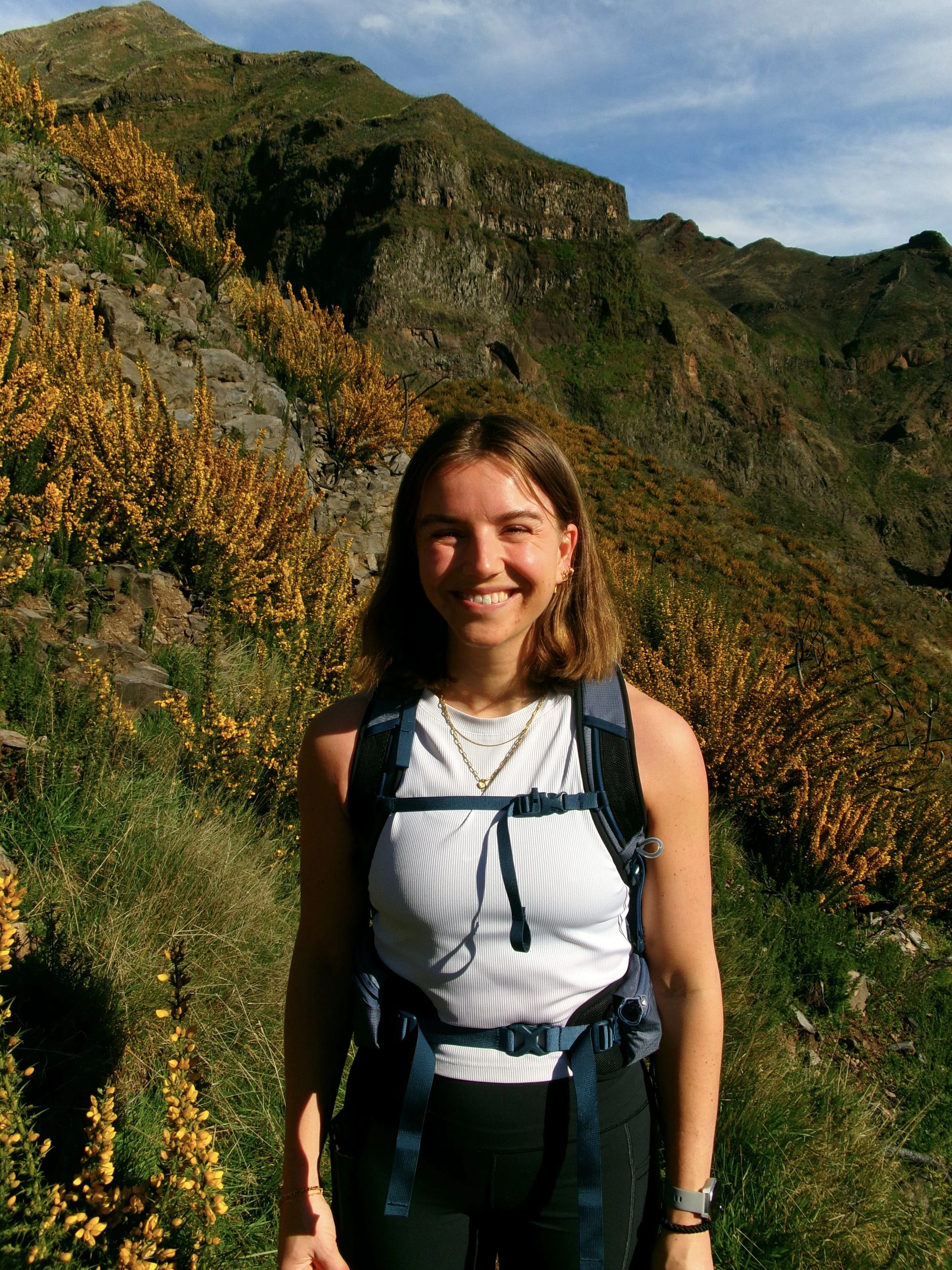 A woman with shoulder-length brown hair smiling outdoors during daytime in front of a mountain landscape with green and rocky terrain and yellow flowering plants.