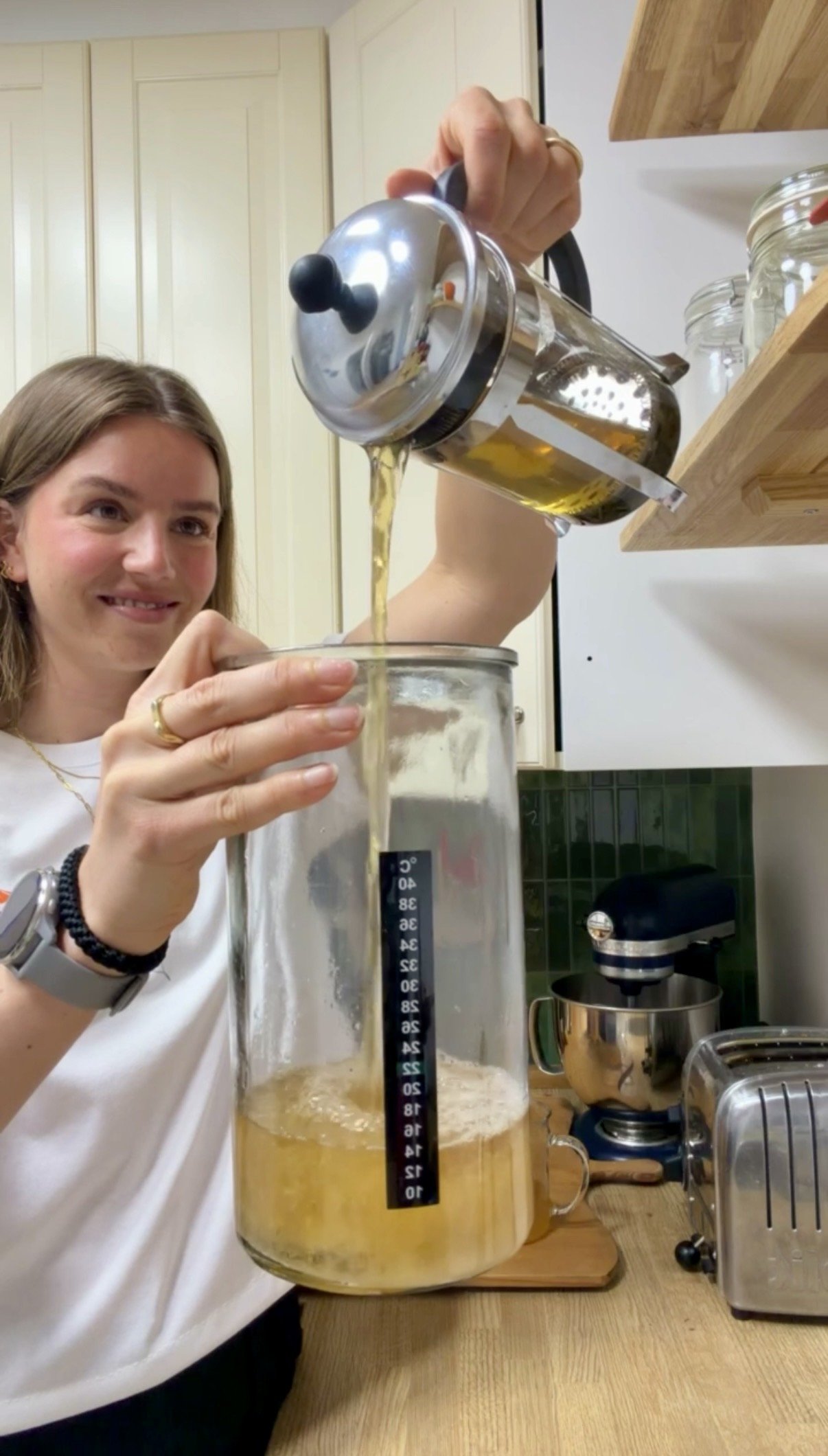A woman pouring liquid into a large measuring beaker in a kitchen, with a smile on her face.