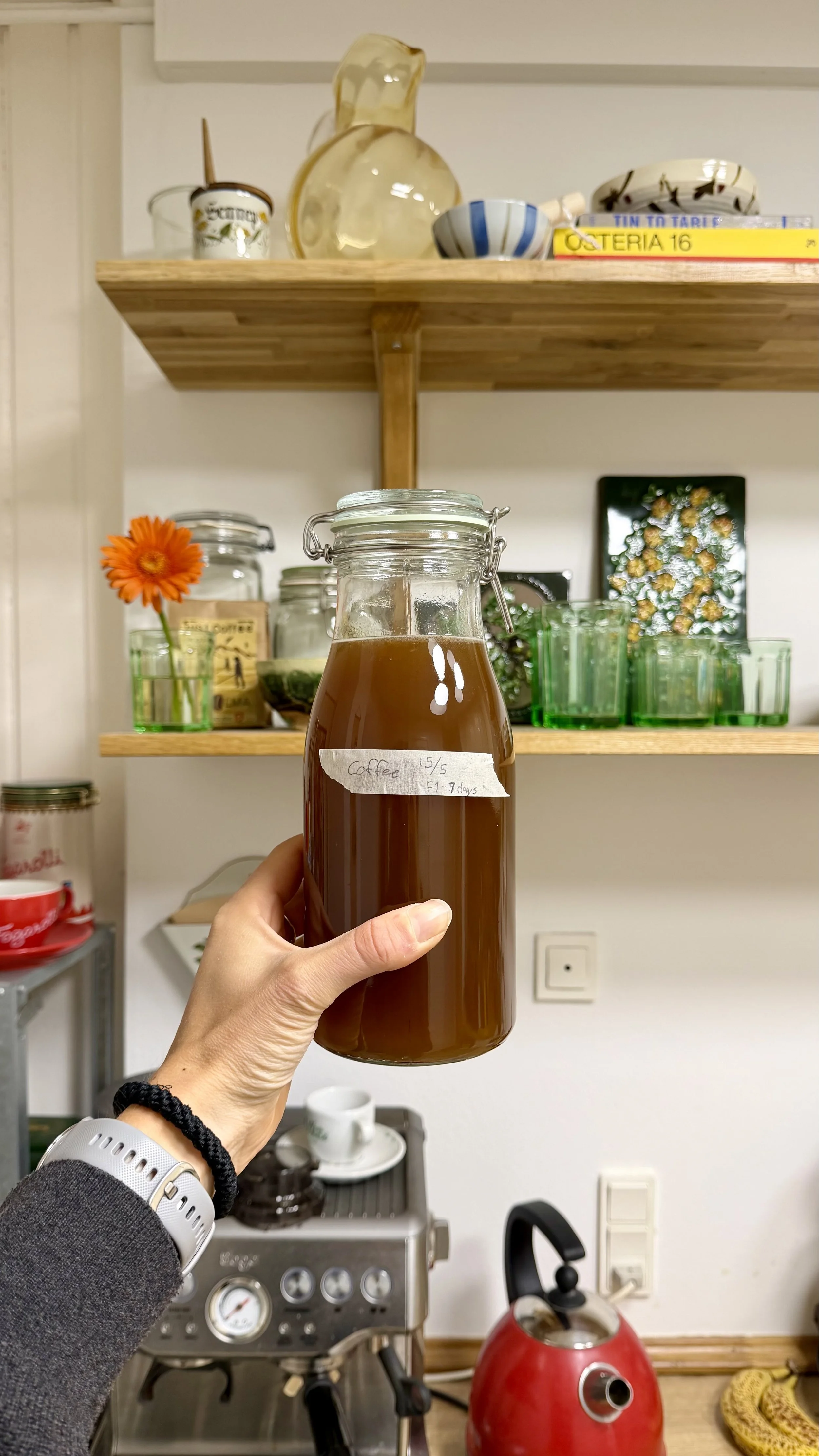A person holding a glass jar labeled 'Coffee 15/5, F4 7 days' filled with dark liquid in a kitchen. Background includes a coffee machine, cups, green glasses, and shelves with bowls and books.