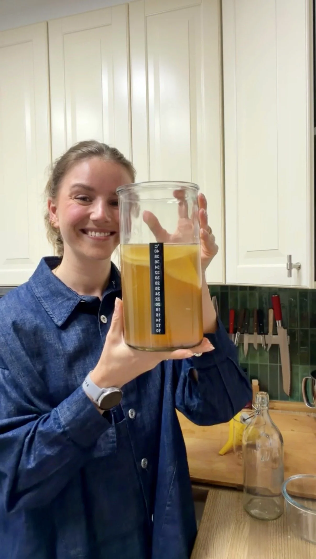 A woman smiling while holding a large measuring container filled with a yellowish liquid in a kitchen.