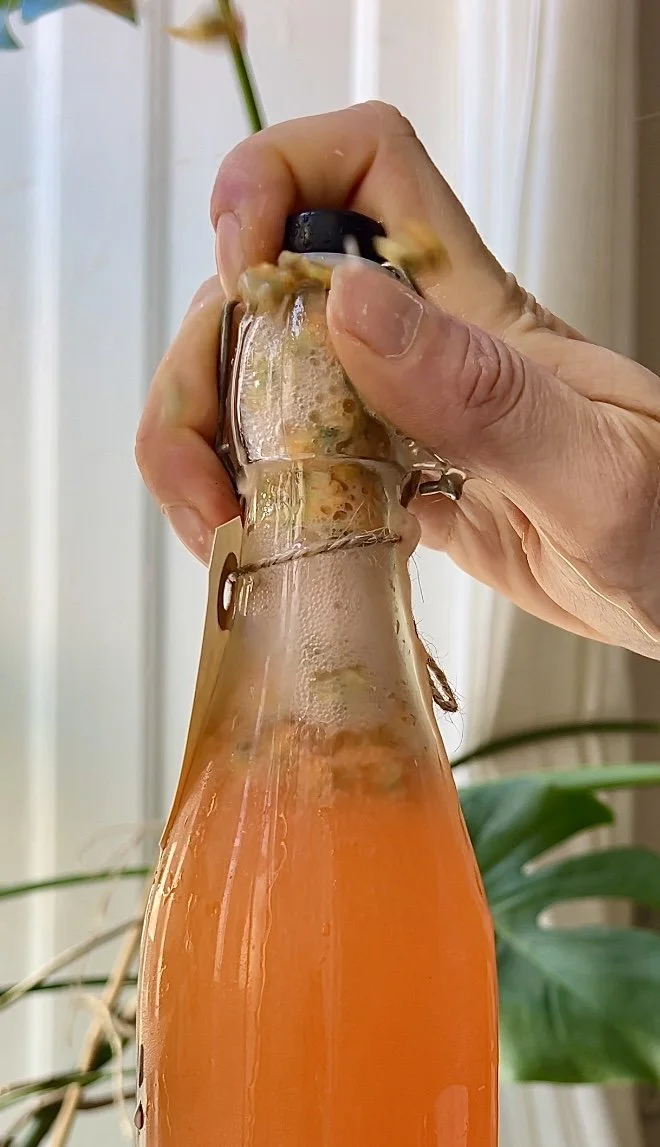 A hand opening a glass bottle with orange-colored beverage inside, with foam and bubbles around the opening, against a background of a white wall and green plants.