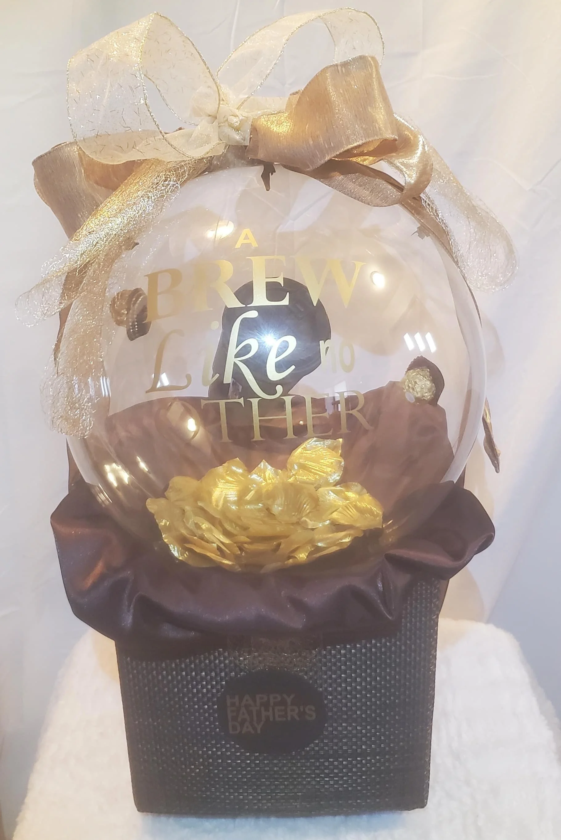 A custom decorative crystal-clear balloon with gold and brown accents, reading 'A BREW LIKE NO OTHER.' The ornament is placed on a dark textured box with a 'HAPPY FATHER'S DAY' adorned with gold lettering and ribbons.