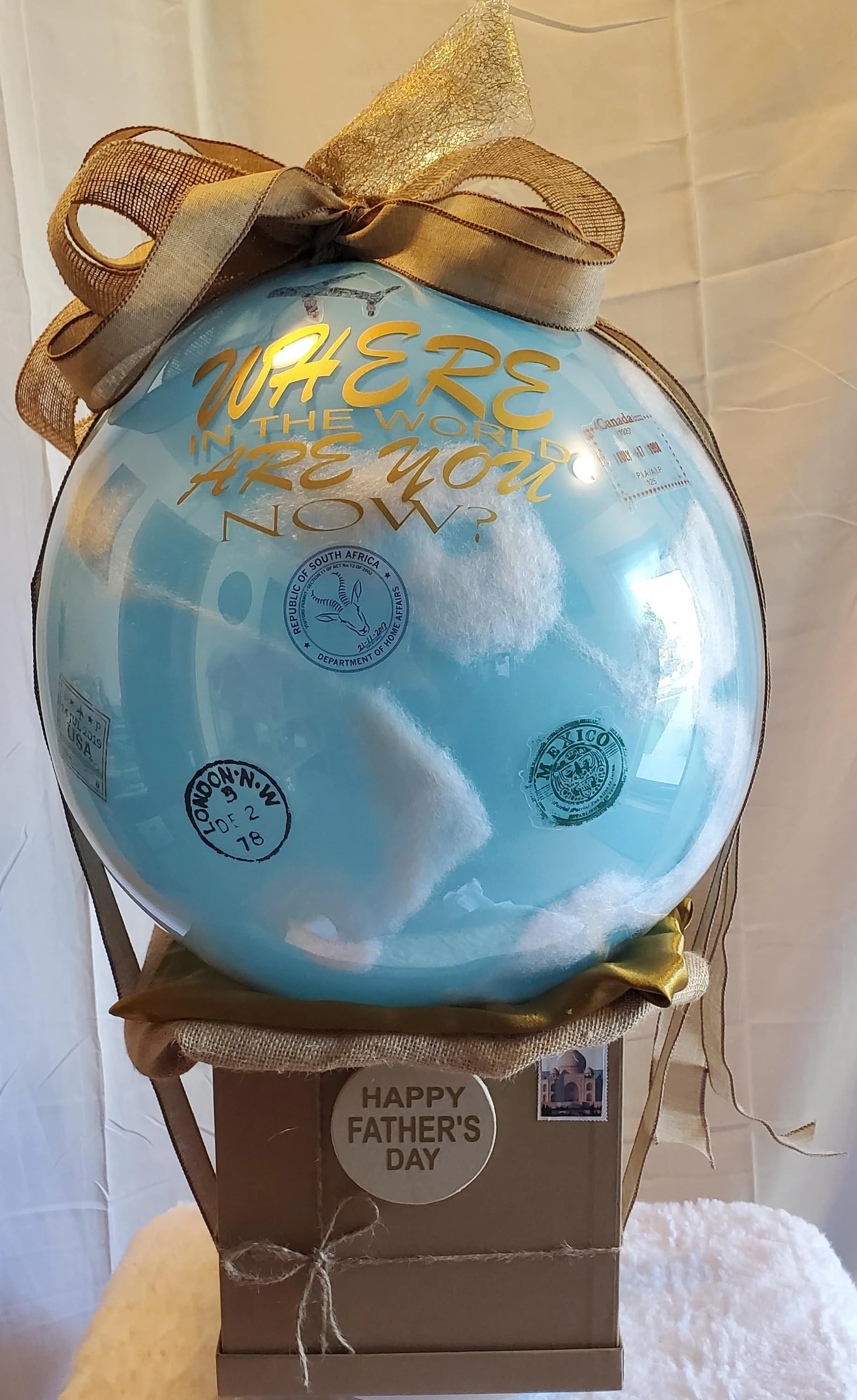 A travel Father’s Day balloon on a gift box designed as a postal package, topped with a large sky designed balloon decorated with gold lettering that reads 'Where in the world are you now?', gold ribbon, and includes international passport stamps.