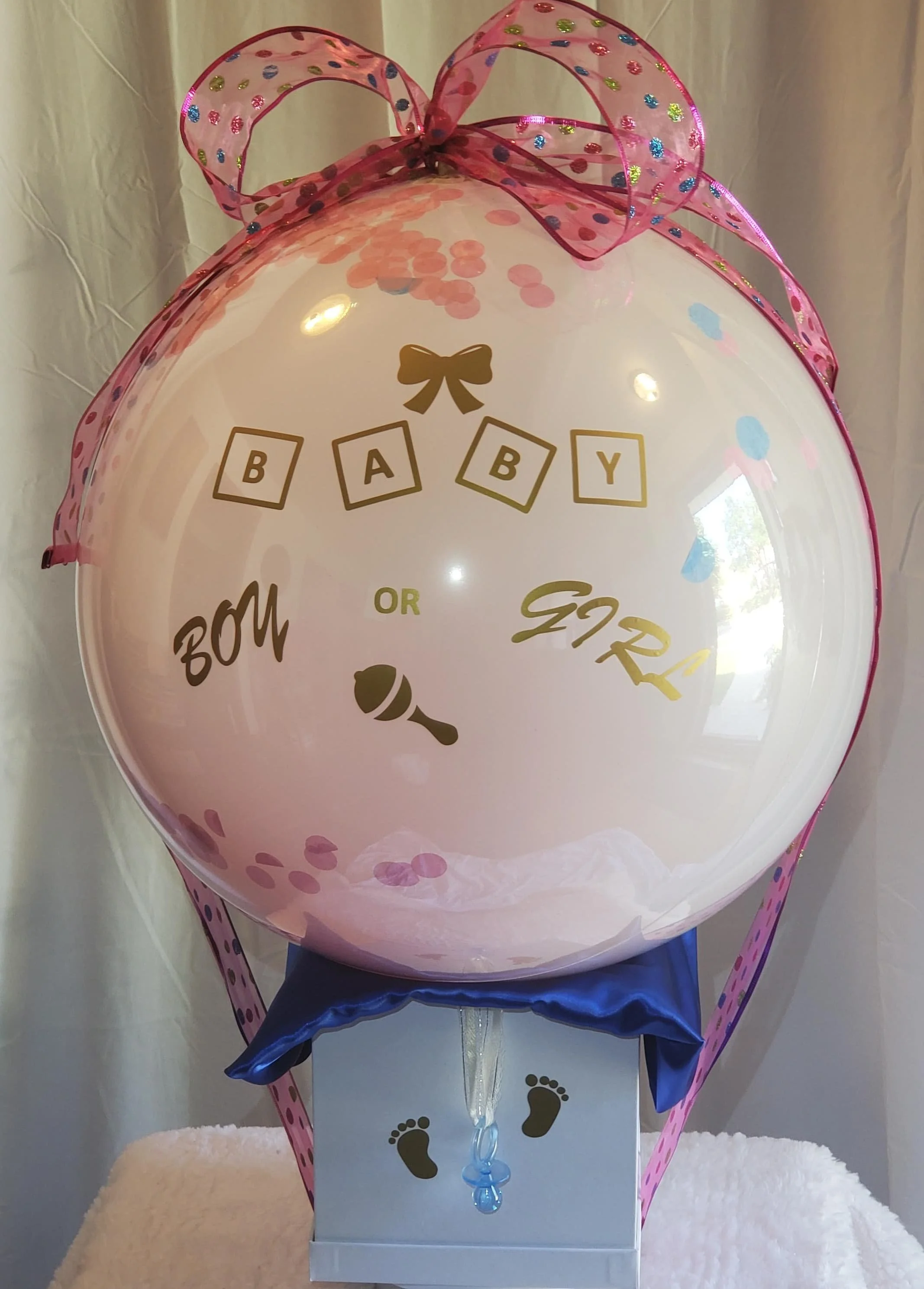 A 'Grand Reveal' pink and blue gender reveal designed balloon with multicolored polka dots, a pink and multicolored sparkling polka dotted ribbon bow on top, and small gold baby footprints on the white gift box.
