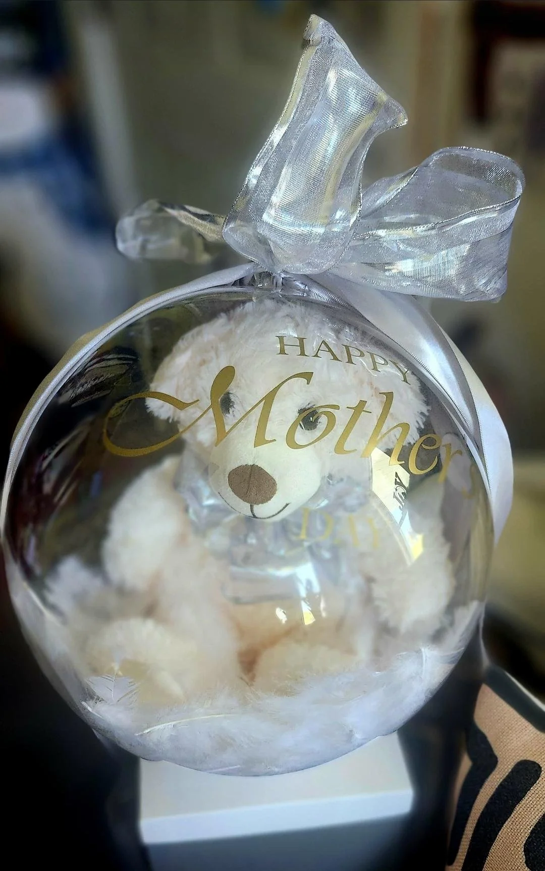 A clear plastic gift bag with a gold 'Happy Mother' message and a teddy bear inside, tied with a large silver ribbon.