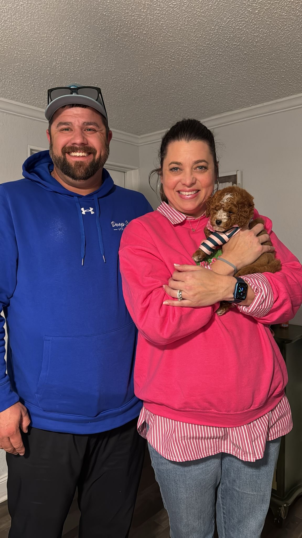A man wearing a blue hoodie and a woman in a pink sweatshirt holding a small puppy, both smiling indoors.