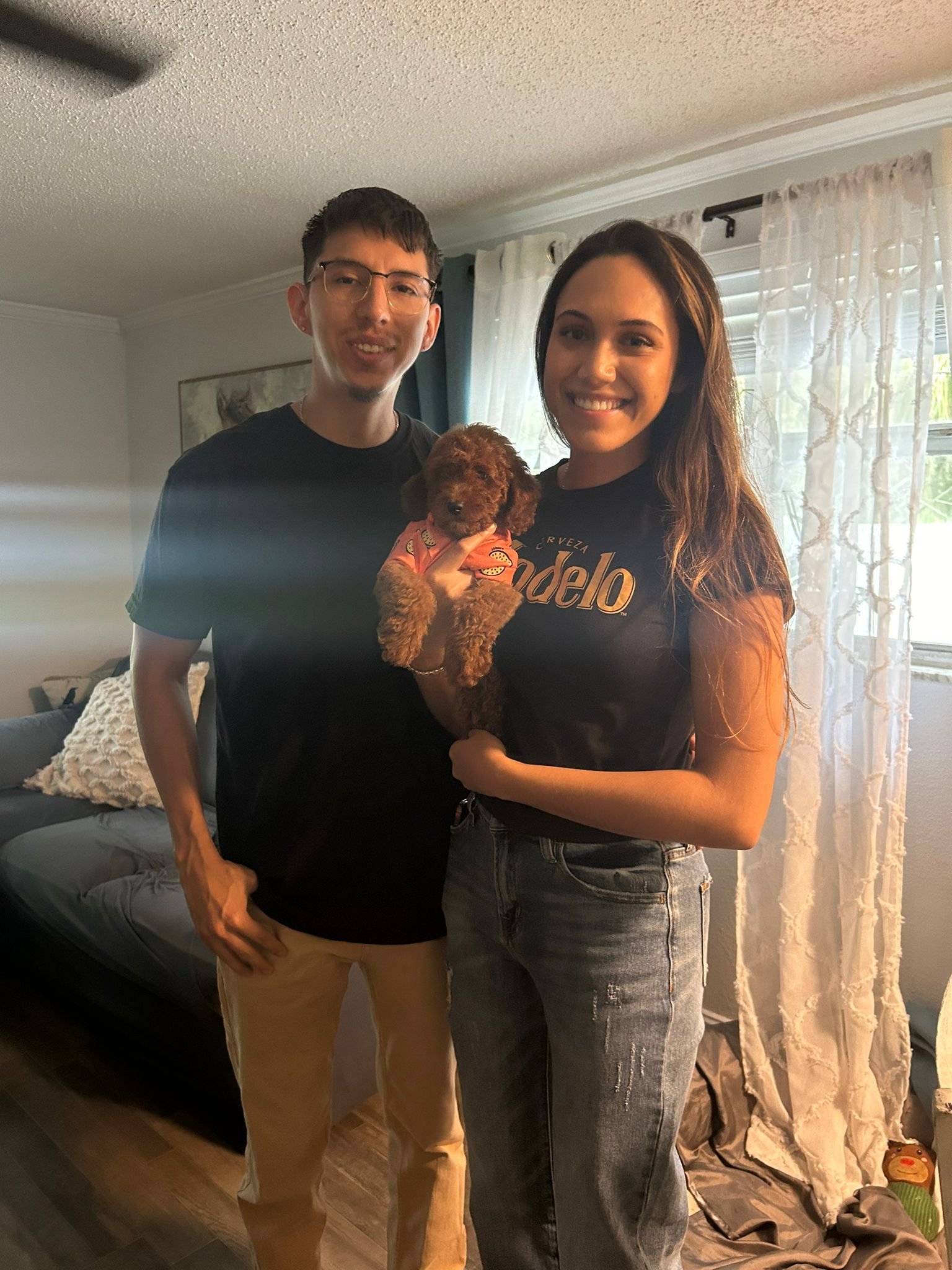 A young man with glasses and a young woman with long hair are standing indoors, smiling at the camera. The woman is holding a small, brown, curly-haired puppy wearing a pink shirt. There is a sofa with a blanket on it, sheer curtains, and a window in