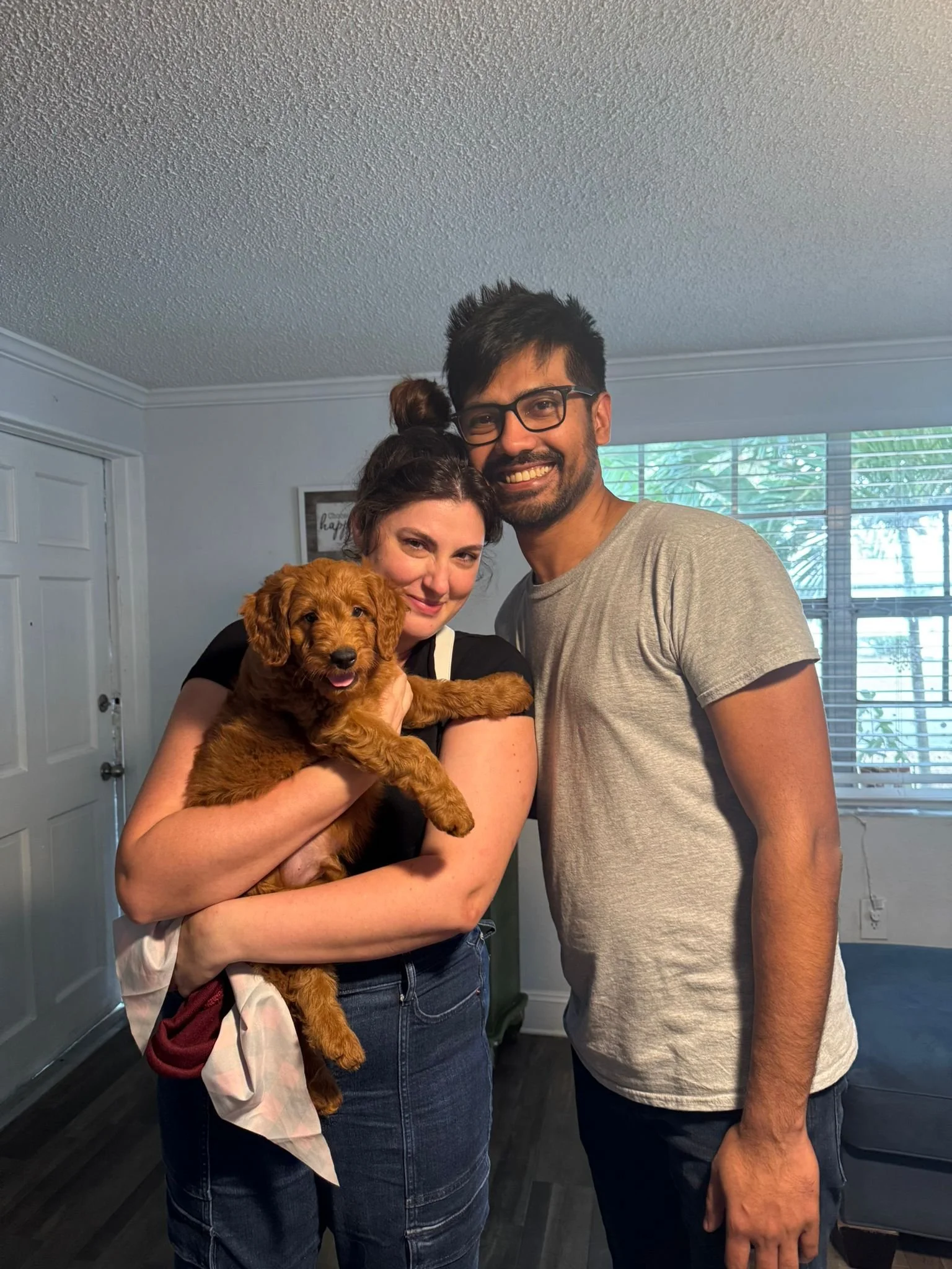 A woman holding a golden retriever puppy and a man with glasses smiling indoors near a window with blinds.