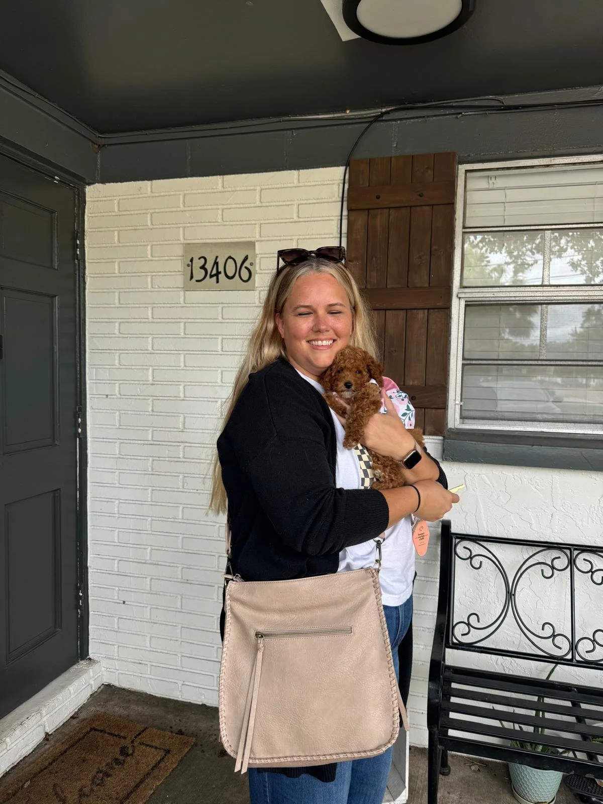 Woman smiling while holding a small curly-haired brown puppy outside a house with white and brick walls, window, and door.