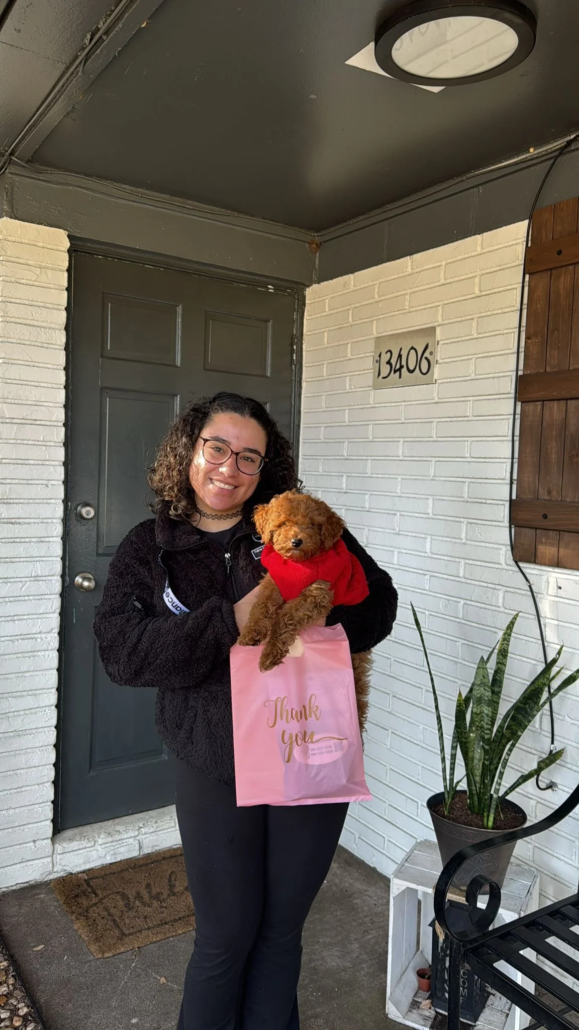 A woman with glasses and curly hair holding a small, brown puppy in a red sweater. She is standing outside a house with a gray door, white brick walls, and a house number 13406. She is holding a pink gift bag with "Thank you" written on it.