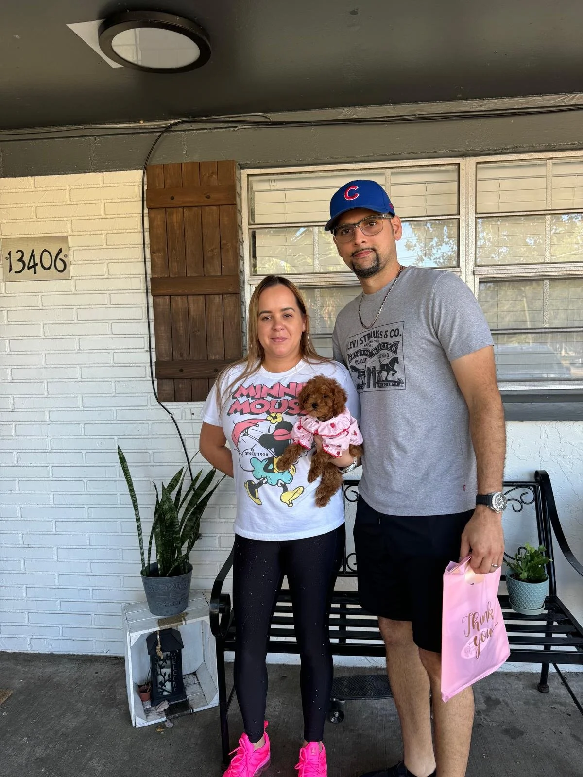 A woman and a man standing on a porch holding a small dog dressed in pink. The woman is wearing a Minnie Mouse t-shirt and black leggings with pink sneakers. The man is wearing a gray t-shirt, black shorts, a blue baseball cap, and glasses, holding a
