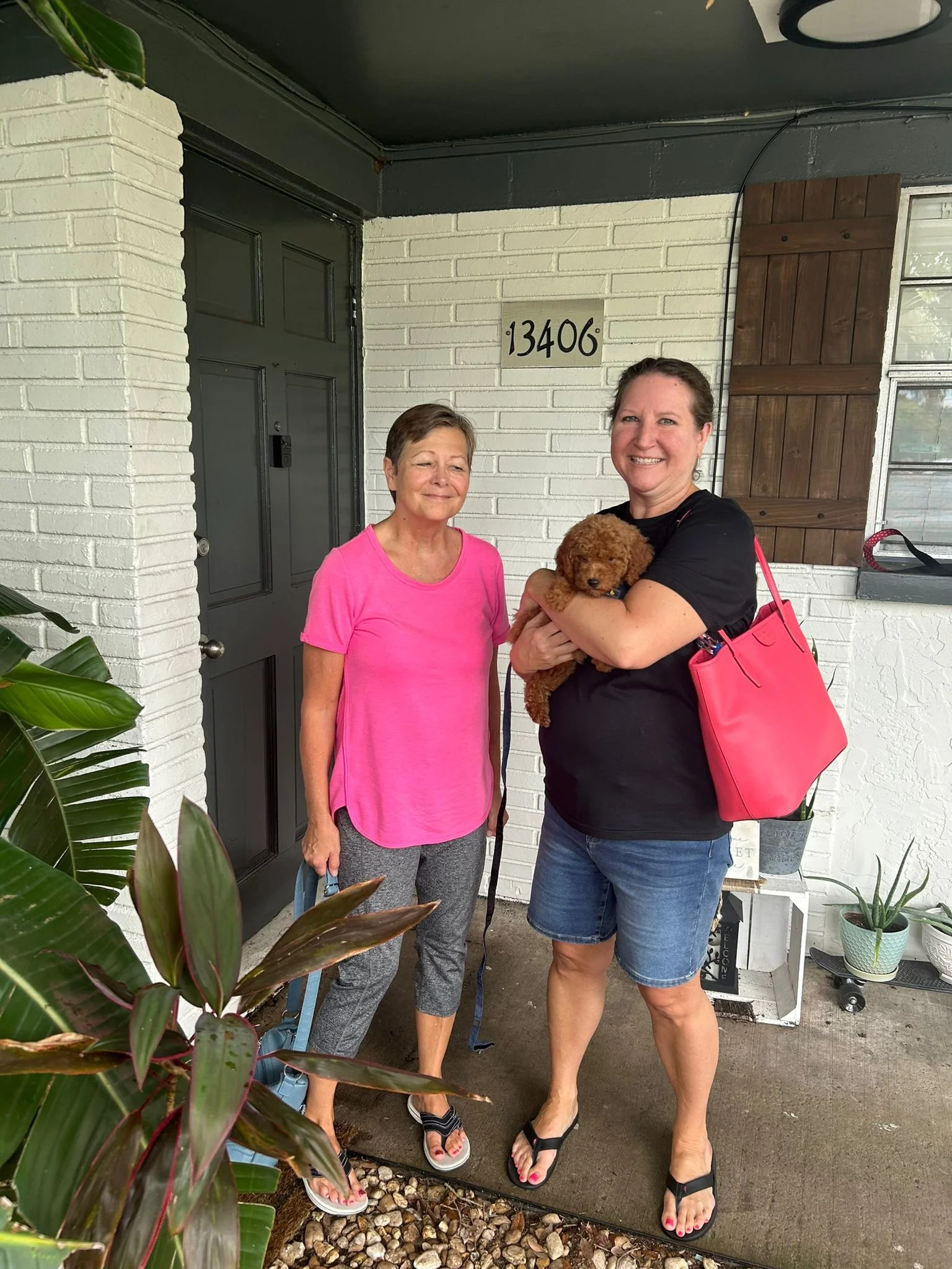 Two women standing outside a house with white brick walls and a house number 13406; one woman is holding a small brown puppy, both are smiling. There are plants and gardening items around.