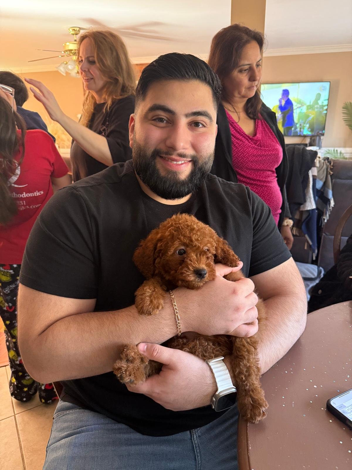 Man with a beard smiling and holding a small brown puppy in a social gathering with women in the background.