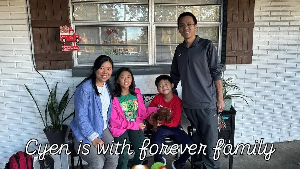 A family of four, including two children, a woman, and a man, pose for a photo on a porch with a white brick wall and window in the background. They are smiling, and the young boy in a red shirt is holding a small brown puppy. There are potted plants