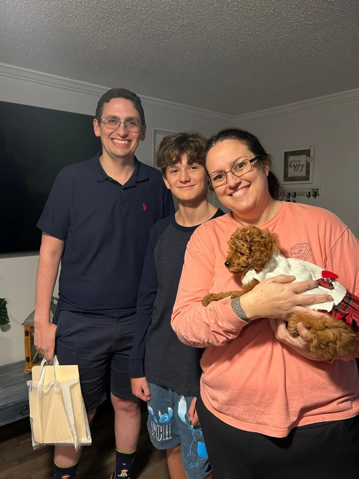 A family of four people and a puppy posing indoors for a photo. All are smiling and standing close together, with the woman holding the puppy.