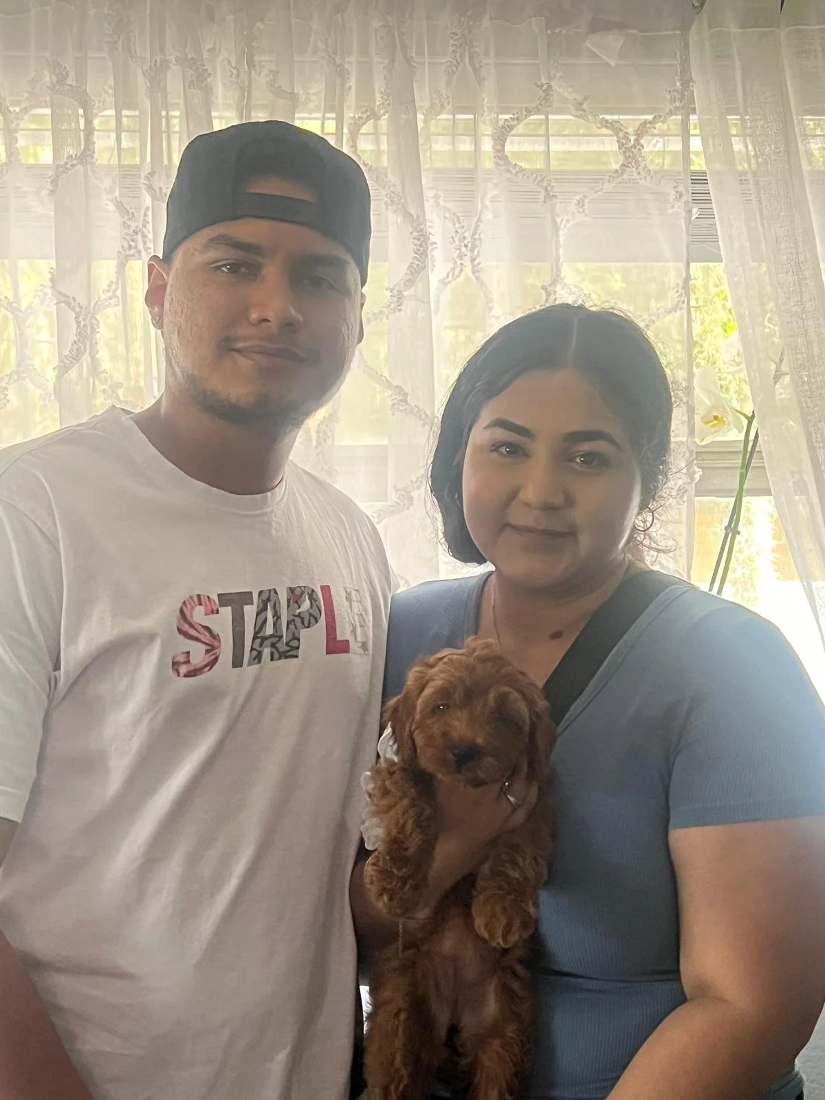 A man in a white T-shirt with the word 'STOP' on it, a woman with dark hair wearing a blue shirt, and a small brown puppy are posing inside a room with lace curtains and a window in the background.