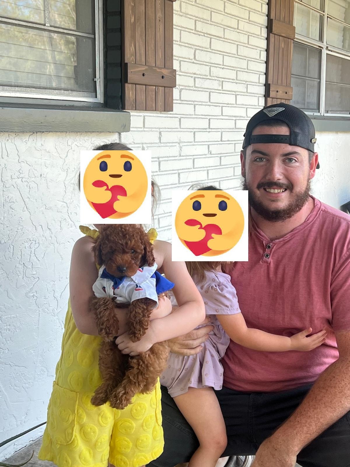 A family photo outdoors on a porch showing a man smiling, two children with emoji faces covering their faces, and a small brown puppy wearing a bandana. The girl in a yellow dress is holding the puppy.